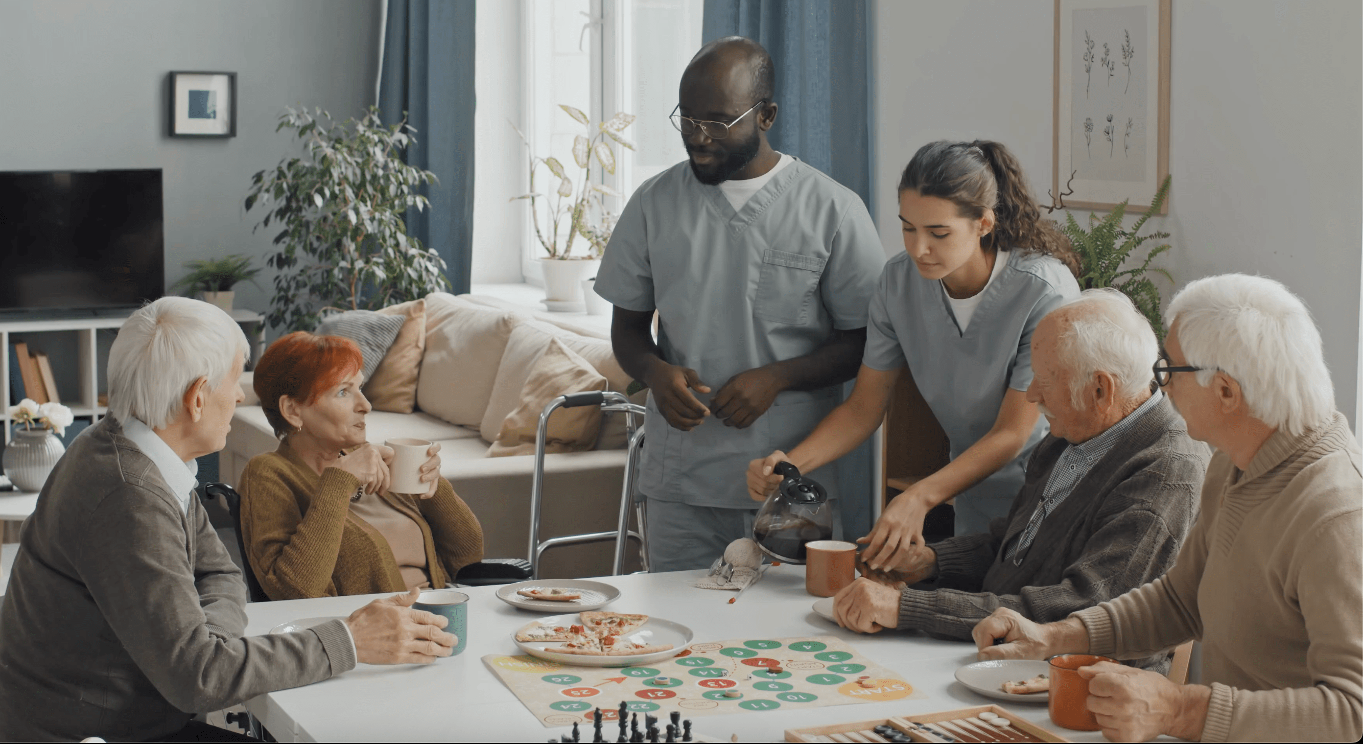 Two nurses pour coffee for a resident with board games on the table