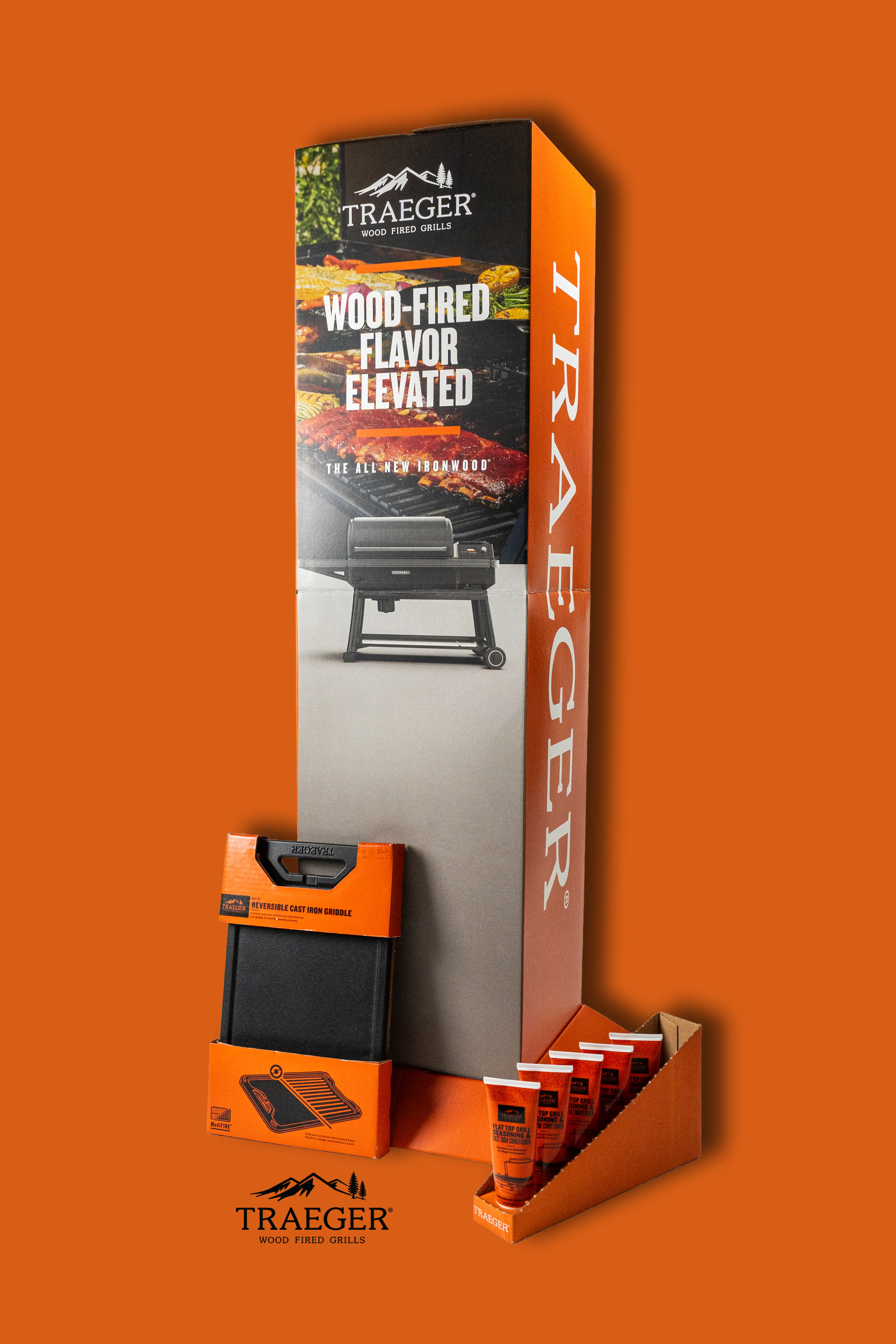 A collection of Traeger products on an orange background, including a reversible cast iron griddle, flat top grill seasoning bottles, and an ironwood grill.