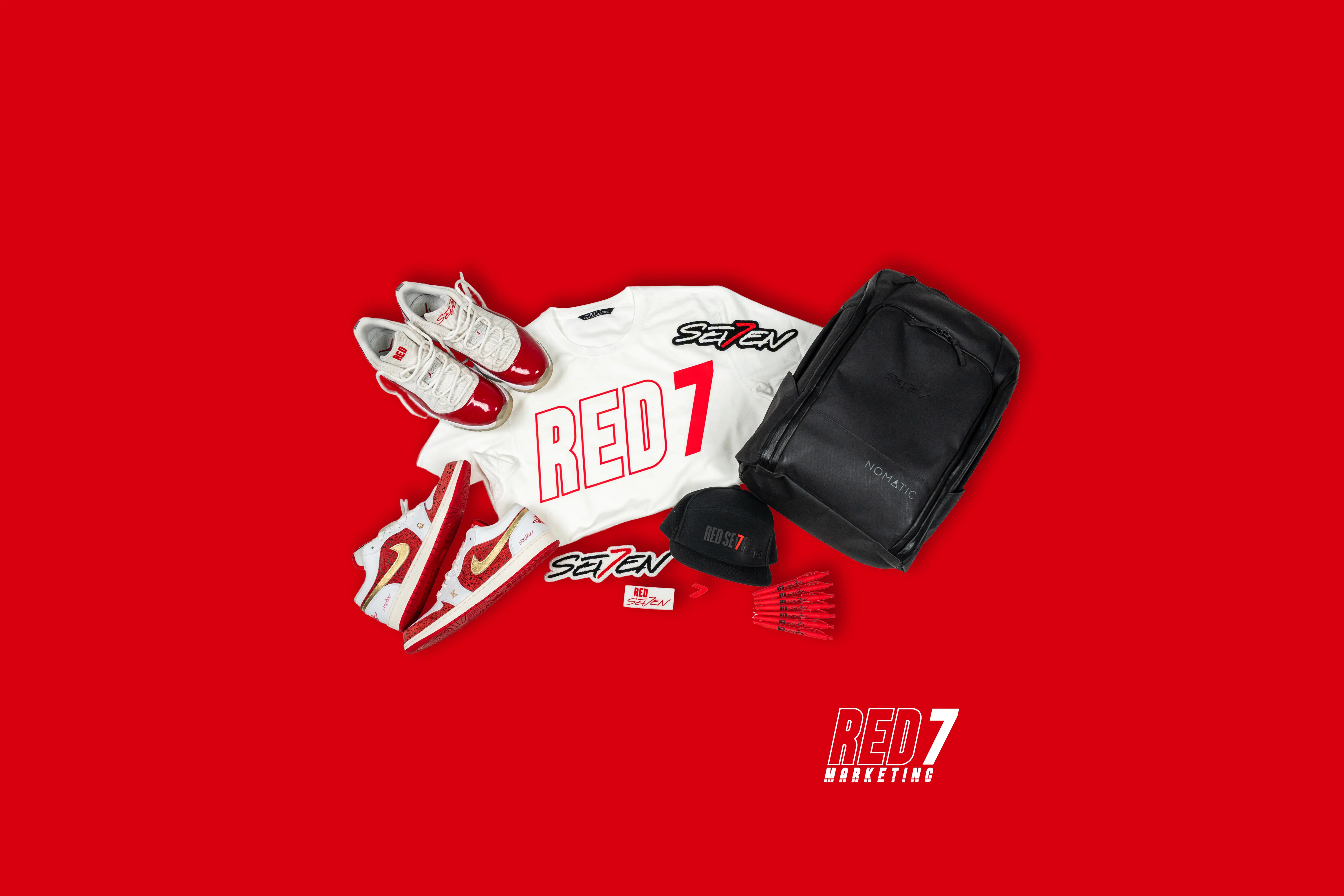 A collection of Red7 branded merch, including shoes, a hat, a tshirt, pens, stickers, and a backpack