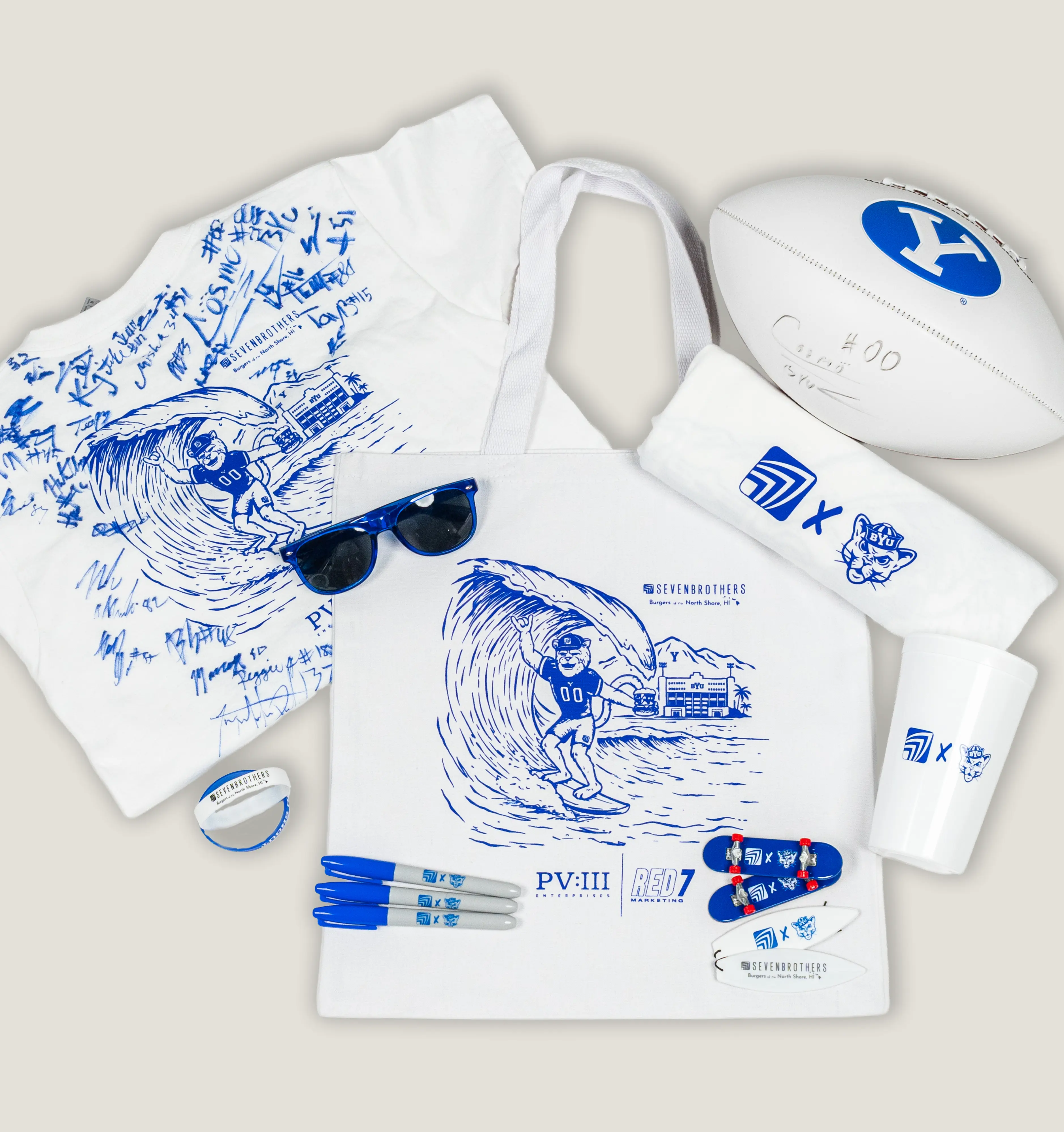 A flatlay of blue and white Seven Brothers x BYU collaboration merch, including a football, travel mug, tshirts, pens, sunglasses, and novelty mini skateboards