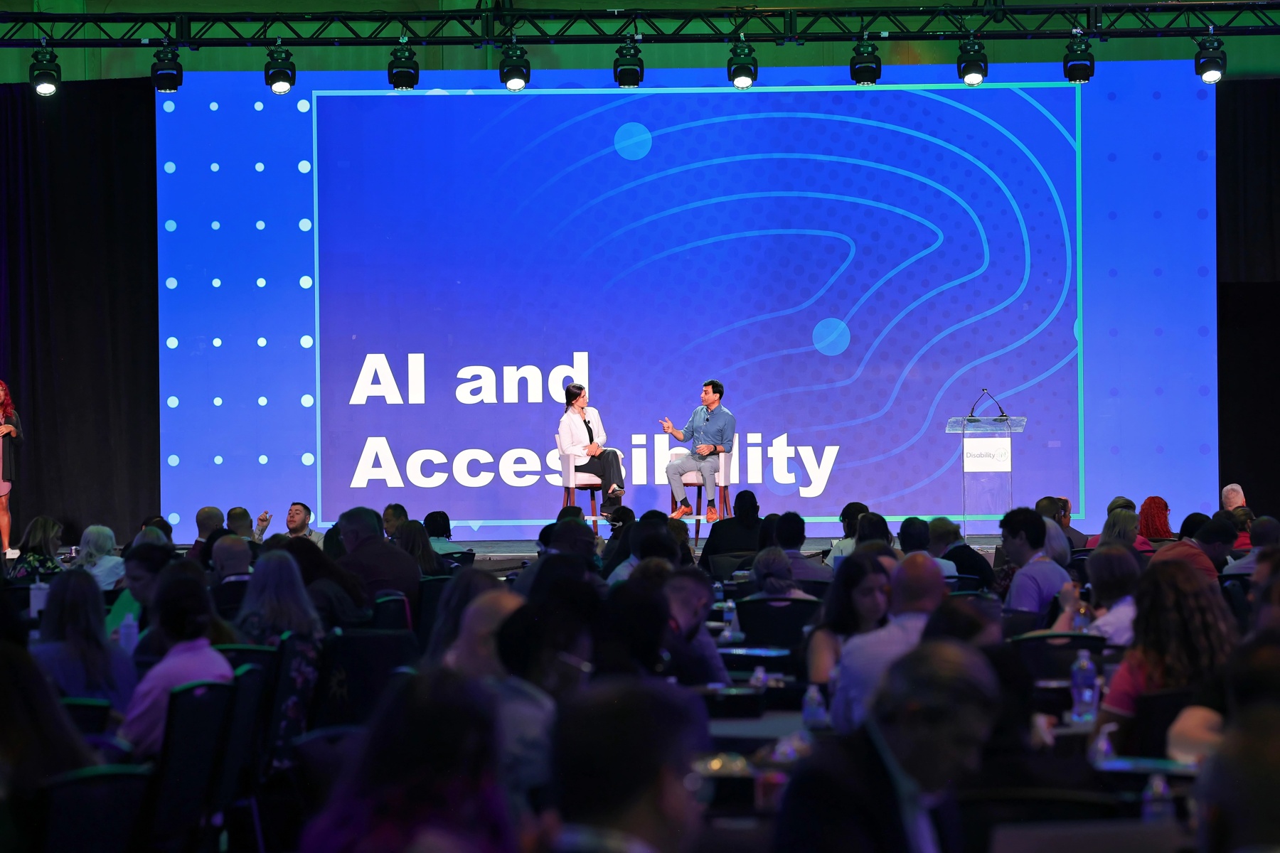 Two professionals speak onstage during a plenary session at the conference. The Audience is full and hundreds of people turn their attention to the stage where "AI and Accessibility" is projected onscreen behind the speakers.