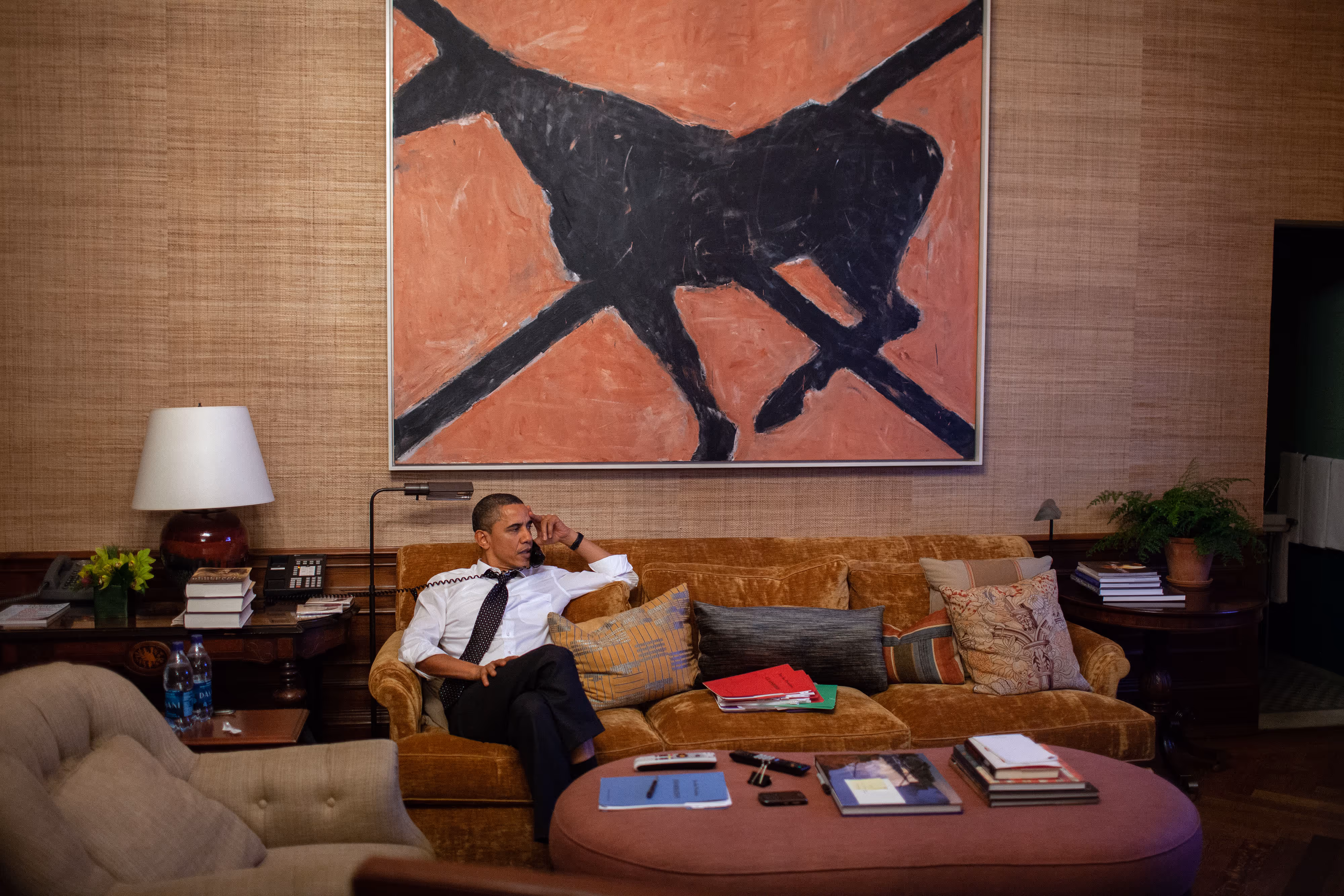 President Barack Obama sits on a sofa talking on the phone beneath a large black horse painting by Susan Rothenberg in a wood-paneled room.