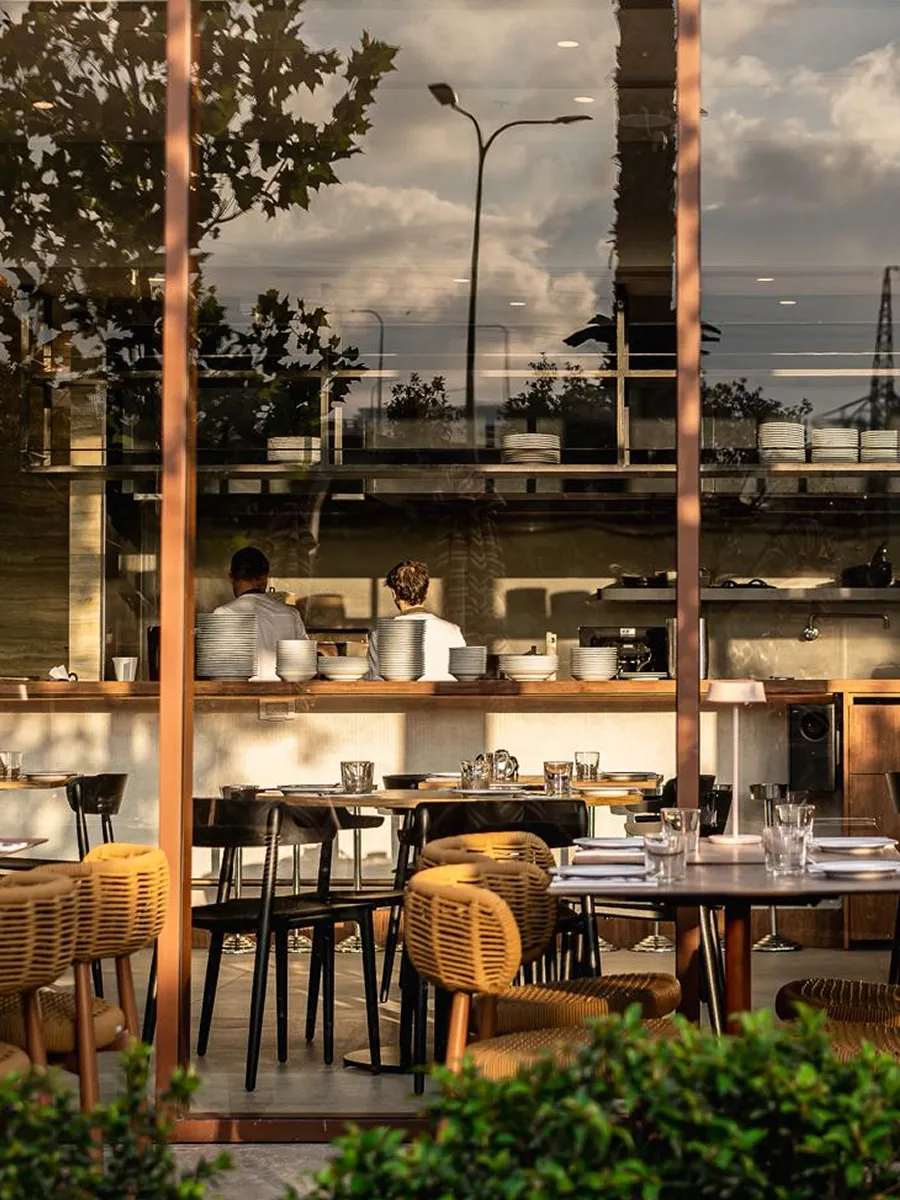 Interior view of a restaurant with stacked plates, tables set with glasses and napkins, and two people sitting at the counter.