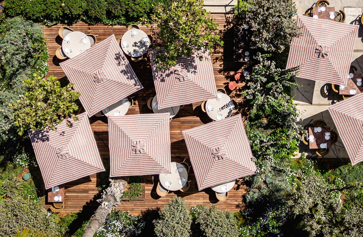 Aerial view of outdoor dining area with round tables, striped square umbrellas, and surrounding greenery.