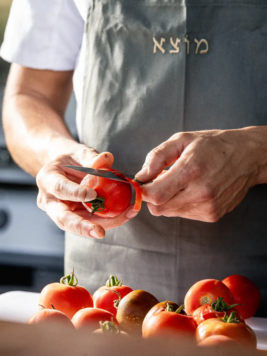 Person peeling a tomato with a knife over a table with various tomatoes.