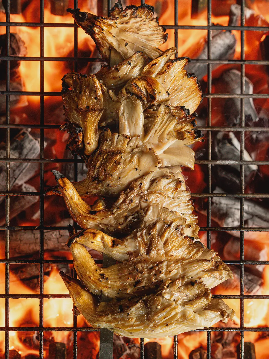 Grilled oyster mushrooms cooking over hot charcoal flames on a barbecue grill.