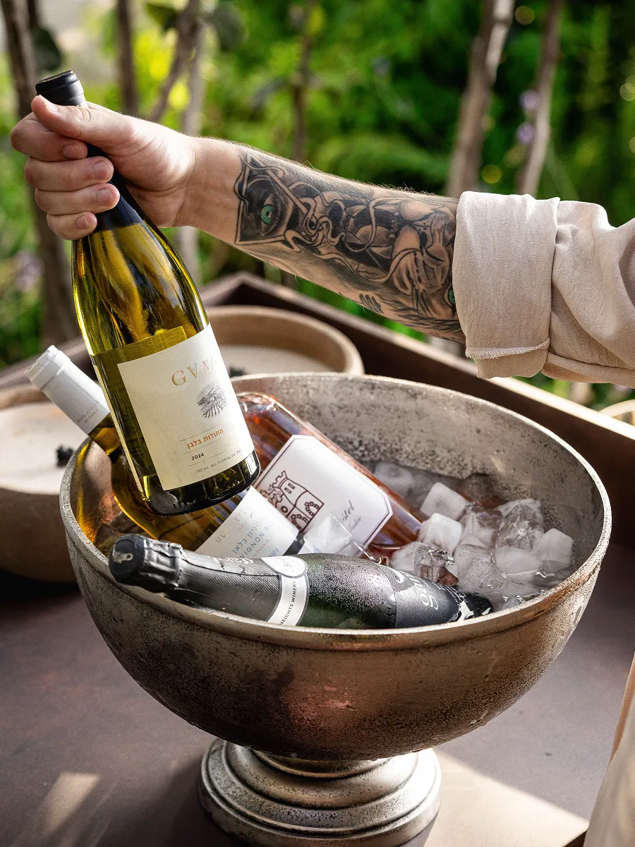 Tattooed arm holding a white wine bottle over a metal bowl filled with ice and assorted wine bottles.