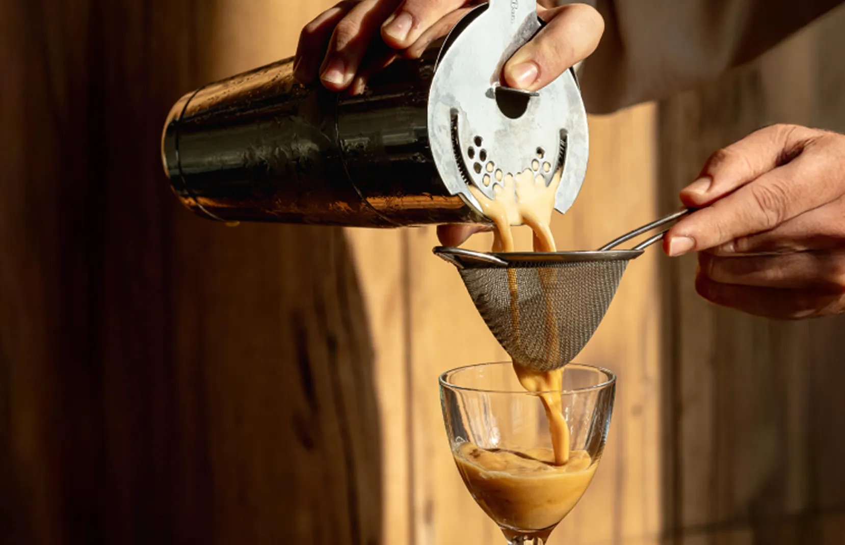 Hands pouring a creamy cocktail through a strainer into a glass.