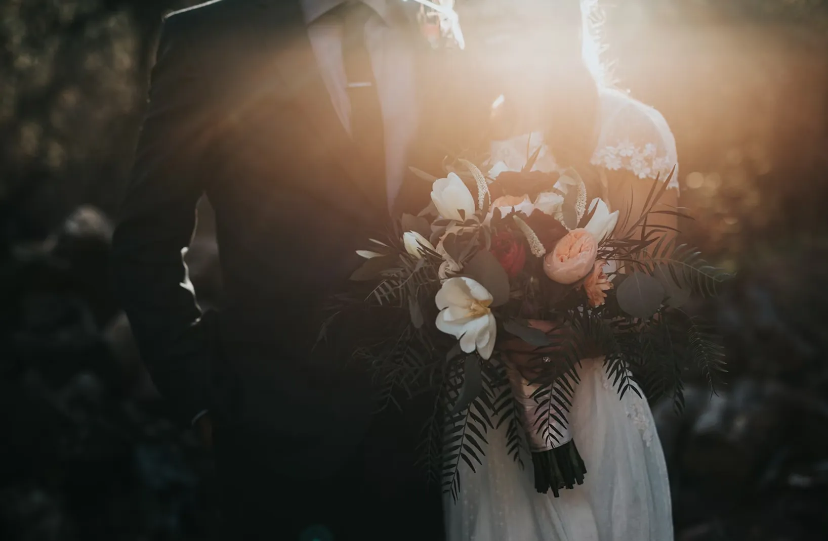 Bride and groom embracing outdoors with the bride holding a bouquet of white, red, and peach flowers in the sunlight.