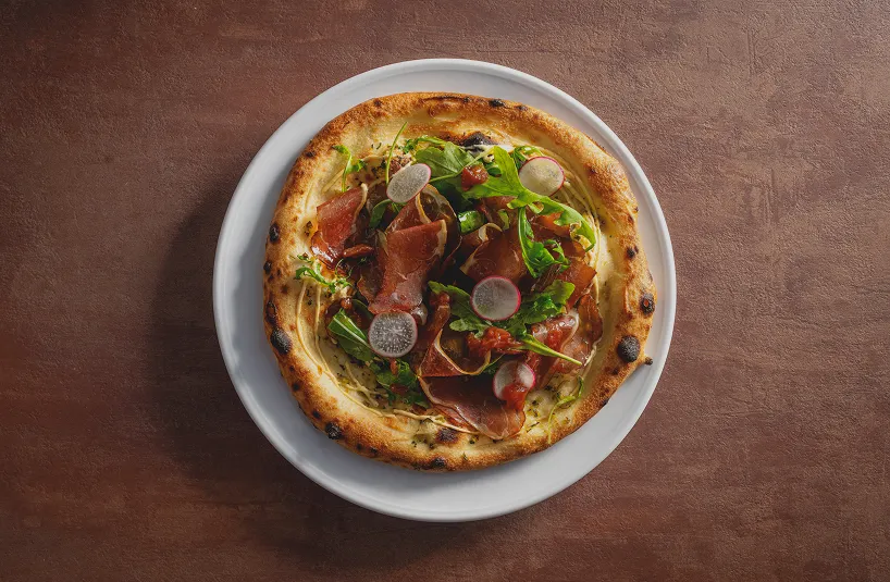 Pizza topped with slices of cured meat, arugula, radish slices, and tomato sauce on a white plate with a brown textured background.