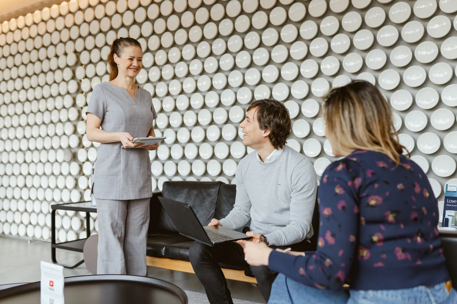 Dental clinic staff member discussing treatment with two seated visitors in a modern lounge area with a textured white wall.