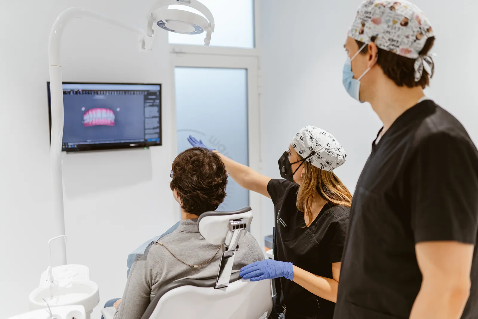 Two masked clinicians and a seated adult look at a dental image on a wall monitor.