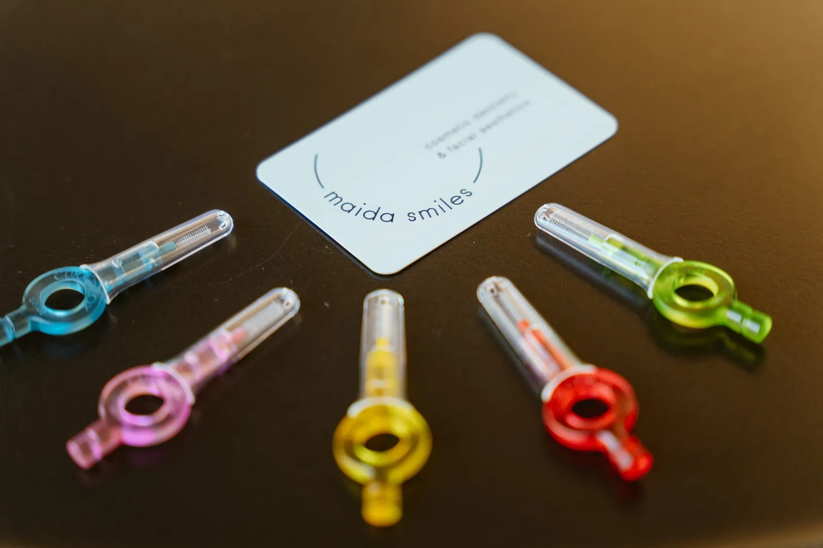 Colourful interdental brushes with clear caps lie beside a Maida Smiles card.