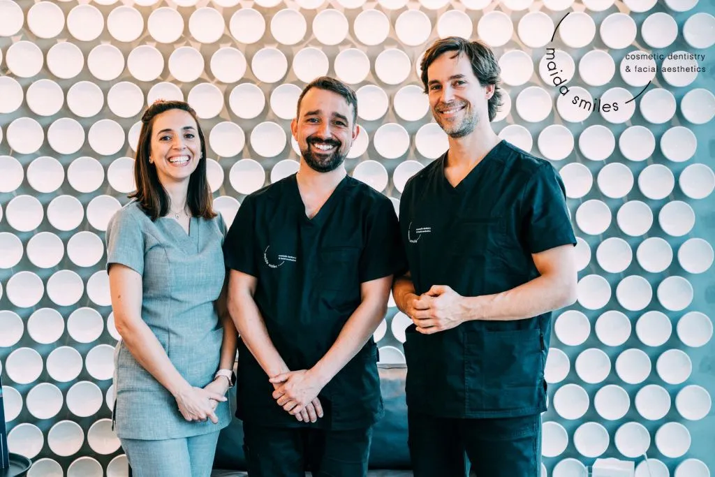 Maida Smiles dental team of three smiling in scrubs in front of the clinic mosaic wall