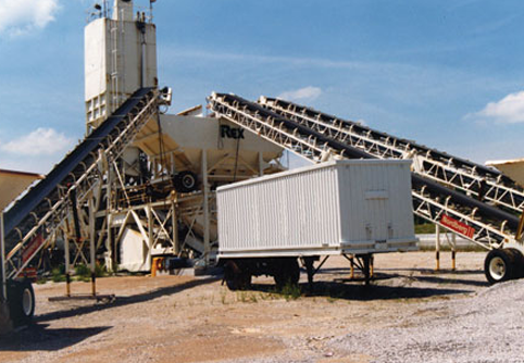 Industrial cement mixing plant with conveyor belts and a white trailer container on gravel ground under a blue sky.