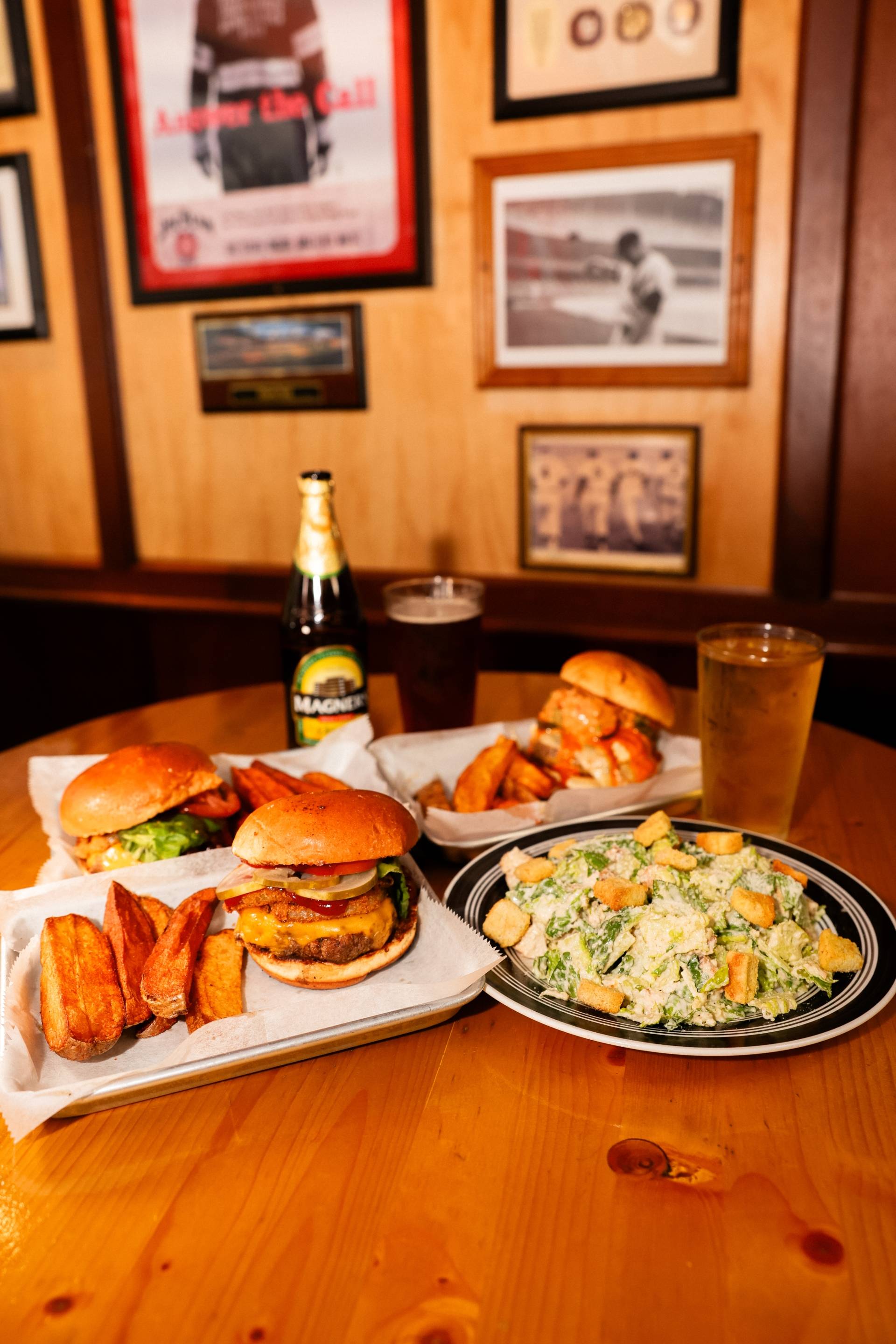 Table with three burgers and potato wedges, a plate of Caesar salad with croutons, a bottle of Magners cider, a glass of dark beer, and a glass of light beer in a pub setting with framed pictures on the wall.
