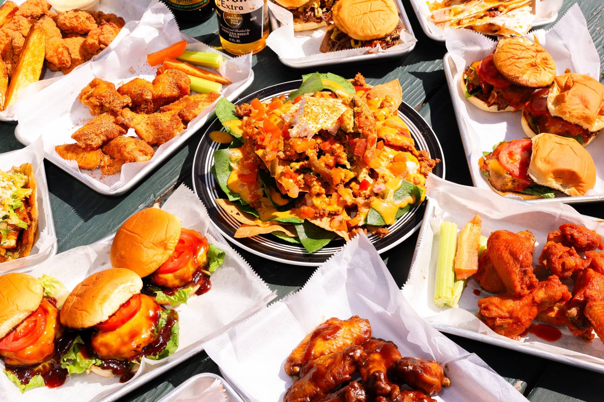 Assorted plates of barbecued chicken wings, sliders, nachos with cheese and toppings, and vegetables on a dark wooden table.