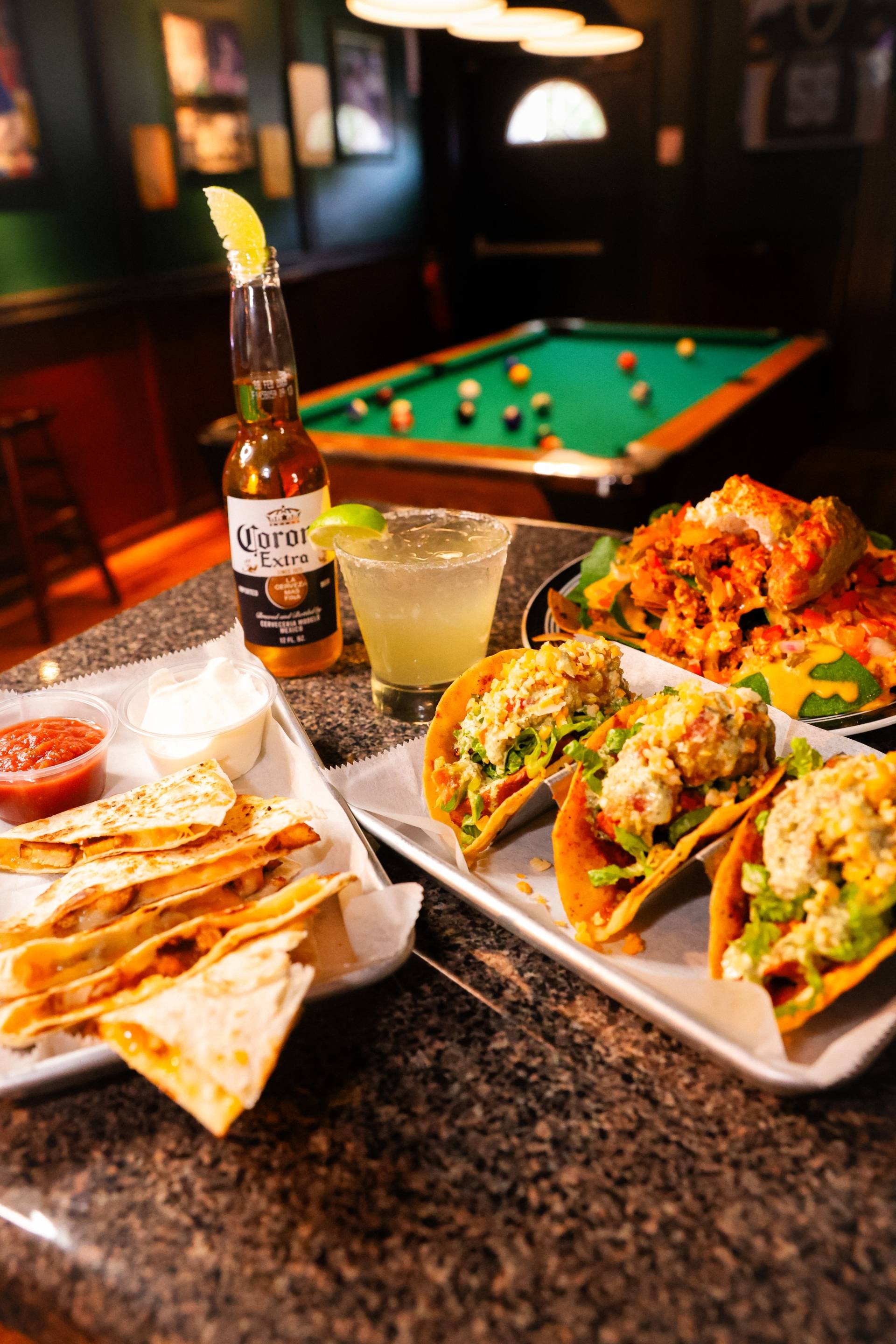 Table with quesadillas, tacos topped with lettuce and cheese, a Corona beer with lime, a margarita glass, and a plate of loaded nachos, with a pool table in the background.