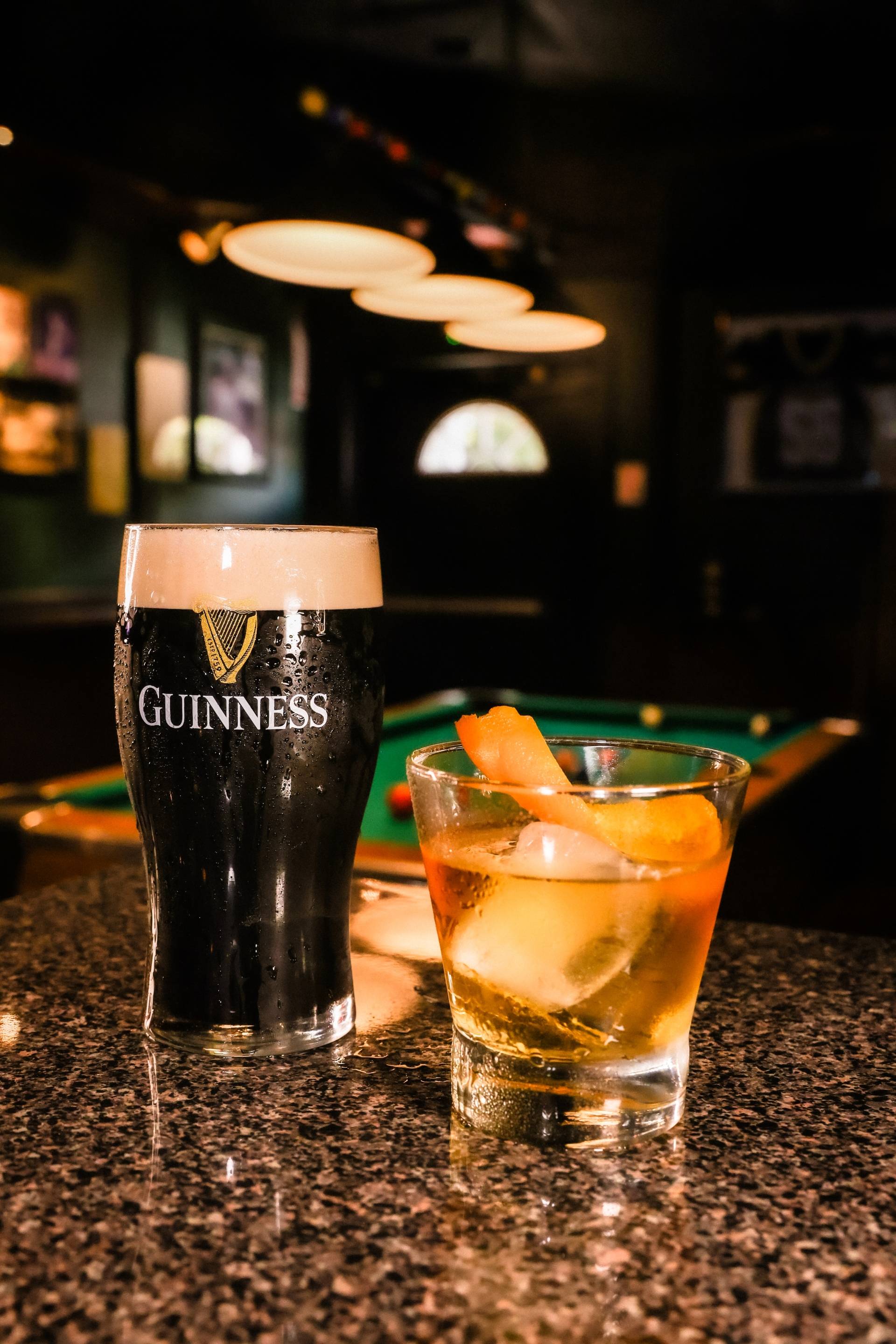 A pint of Guinness beer and a cocktail with ice and orange peel on a bar counter with a pool table in the background.