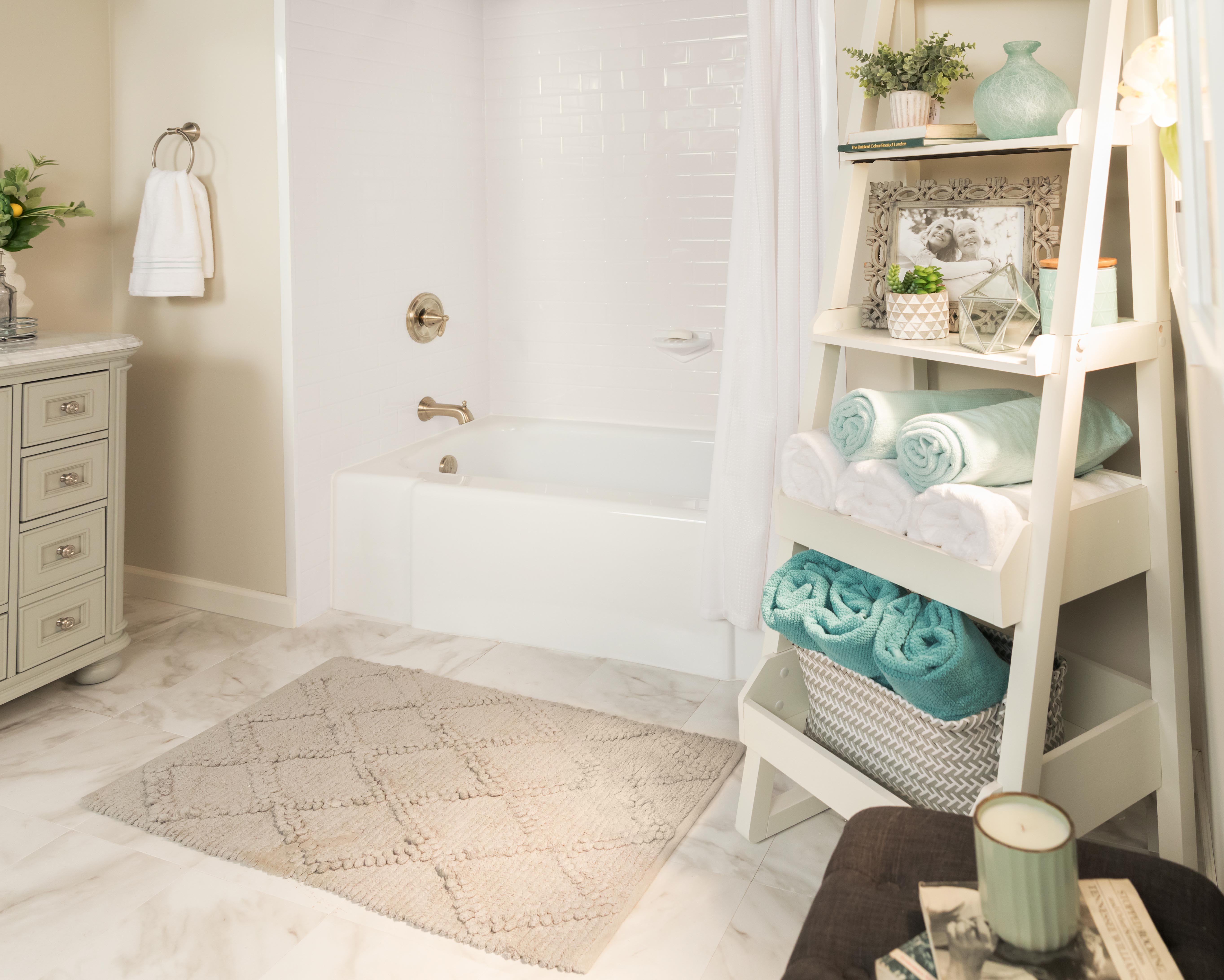 Elegant and cozy bathroom with a soft neutral color palette, featuring a white bathtub, brushed gold fixtures, and marble tile flooring. A tiered ladder shelf holds rolled towels, decorative plants, and framed photos, adding a warm, personalized touch to the serene space.
