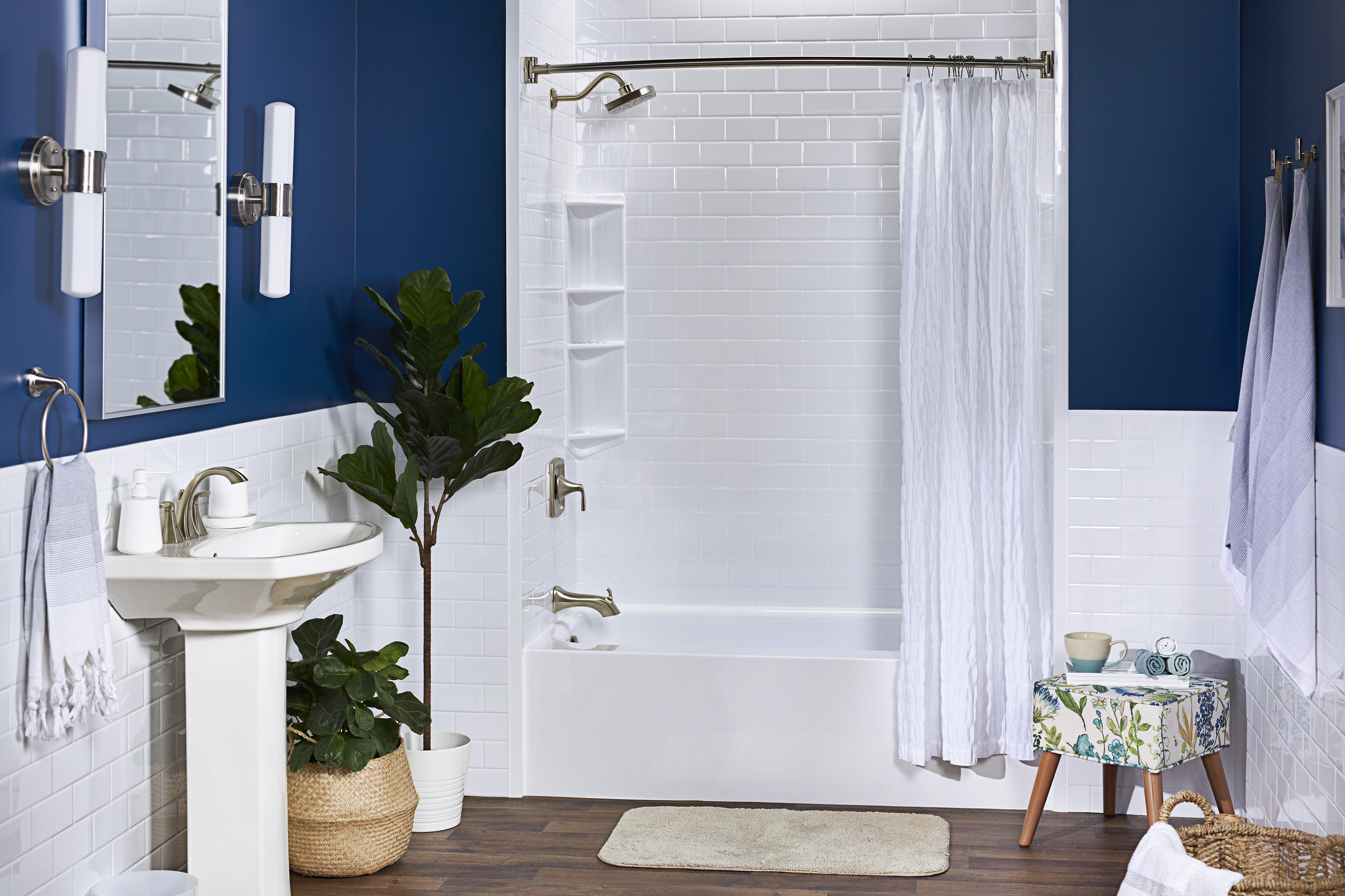 Modern bathroom with a deep blue and white color palette, featuring a white subway-tile shower, brushed nickel fixtures, and wood-style flooring. A pedestal sink, potted plants, and a floral upholstered stool add warmth and character to the space.