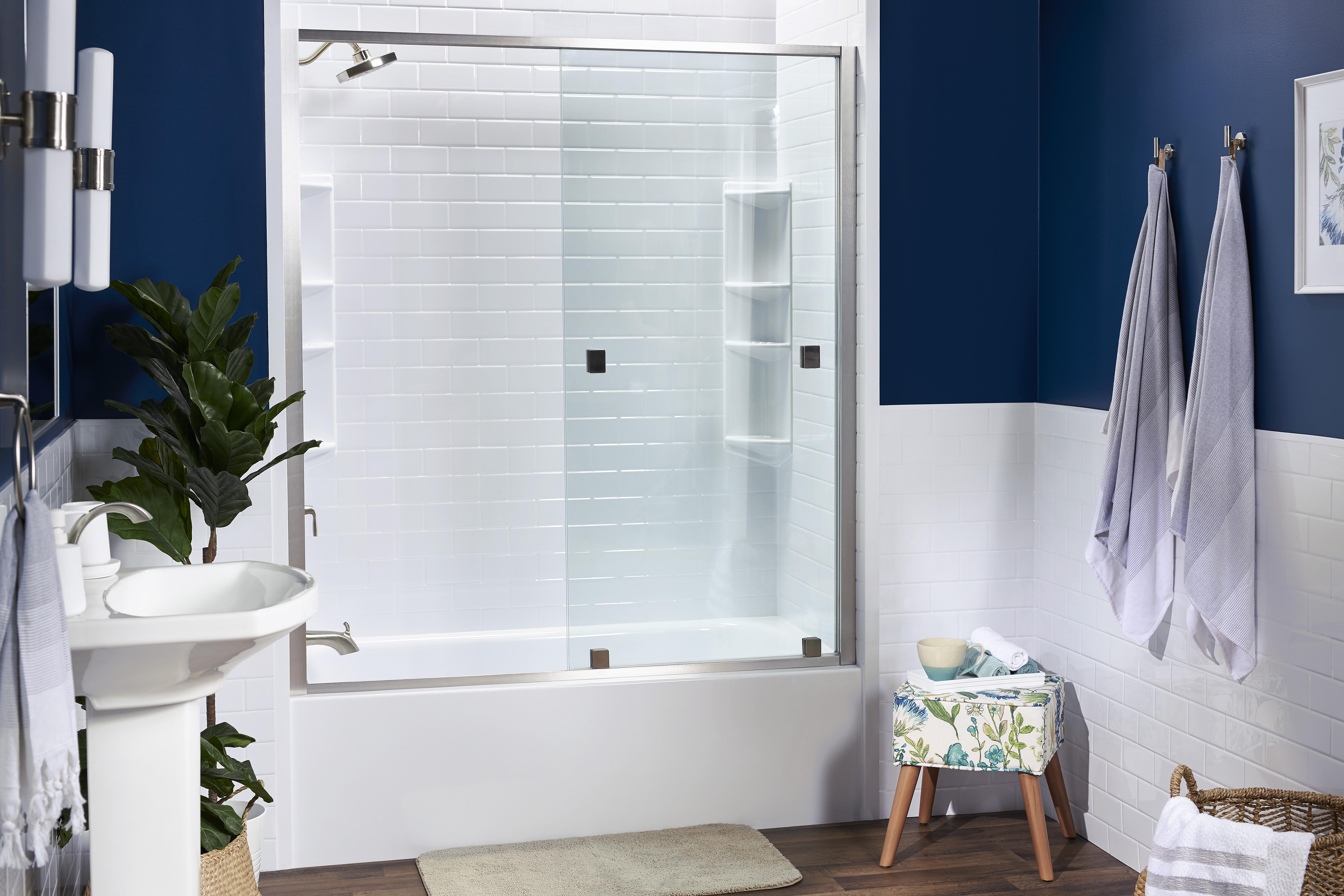 Contemporary bathroom with navy and white walls, featuring a sleek glass shower door over a white bathtub with subway tile surround. The space includes a pedestal sink, potted plant, and a floral-patterned stool, creating a clean, modern, and inviting atmosphere.