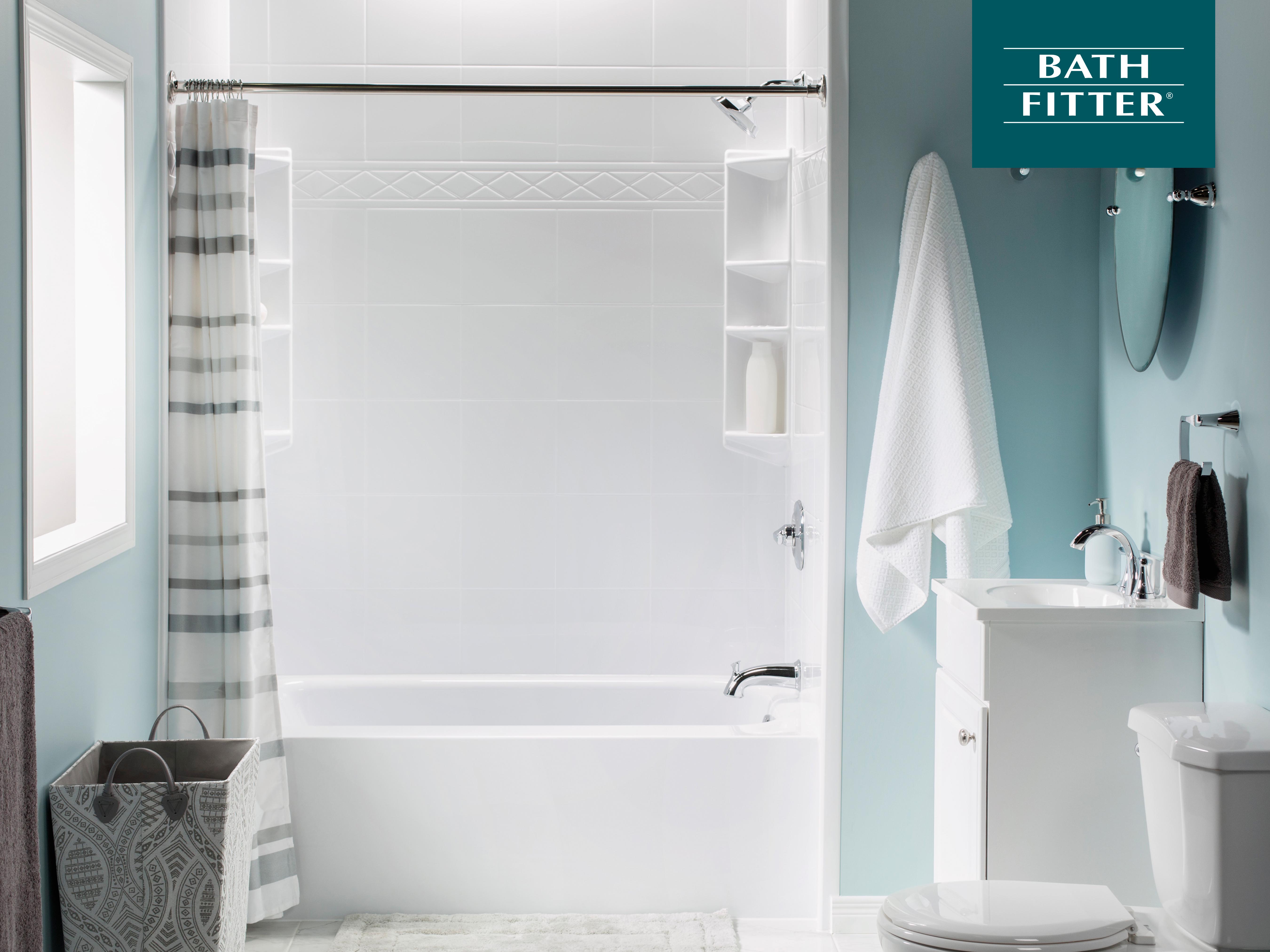 Bright and modern bathroom with a white tub-shower combo featuring built-in corner shelves and a sleek chrome showerhead. A striped shower curtain hangs on a chrome rod, while a compact white vanity with a mirror and towel hooks complements the soft blue walls. A gray laundry basket and bath mat complete the space.