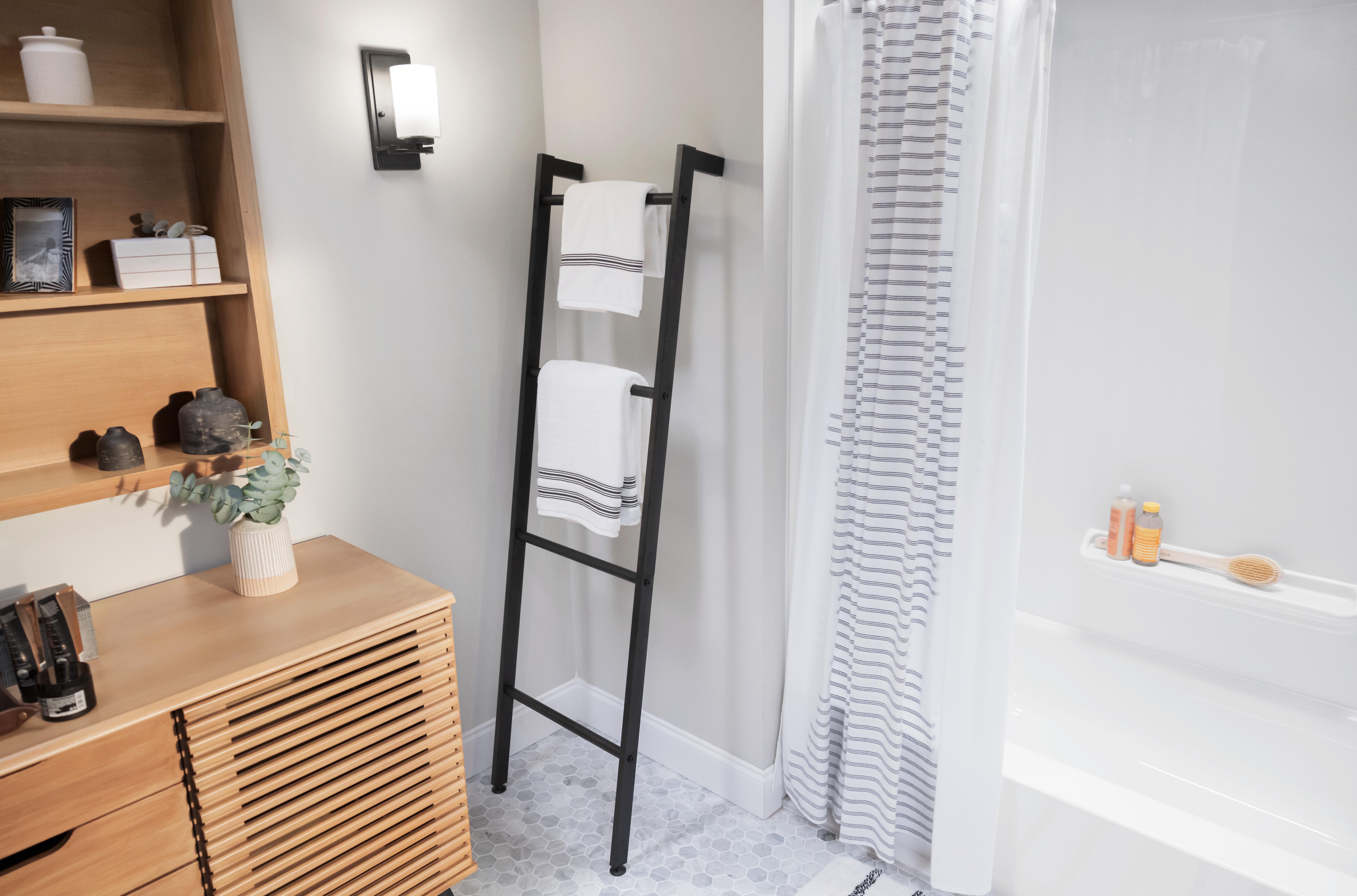 Modern bathroom with a cozy, minimalist design featuring a black ladder towel rack holding white towels with striped accents. A white shower with a striped curtain includes bath products and a brush on a built-in ledge. The space is accented by light gray walls, hexagonal tile flooring, and natural wood furniture with potted greenery and decorative accessories.