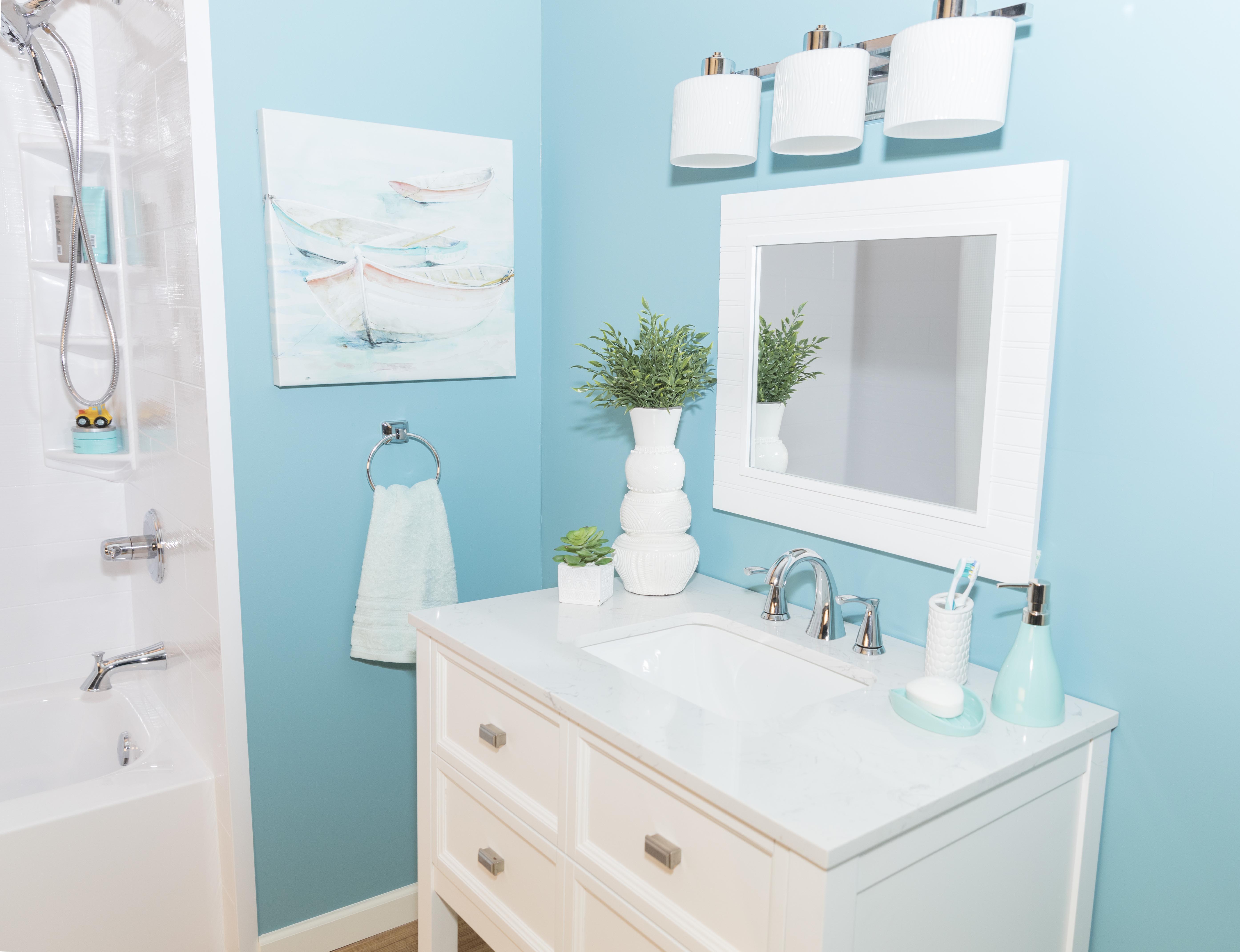 Bright and cheerful bathroom with turquoise blue walls and a white vanity with a marble countertop. The vanity features a modern faucet, soap dispenser, toothbrush holder, and small decorative plants. Above it hangs a white-framed mirror with a three-light chrome fixture. To the left, a bathtub with white tile and built-in shelves is visible, along with a wall art piece depicting pastel-colored rowboats. The space is clean, coastal, and inviting