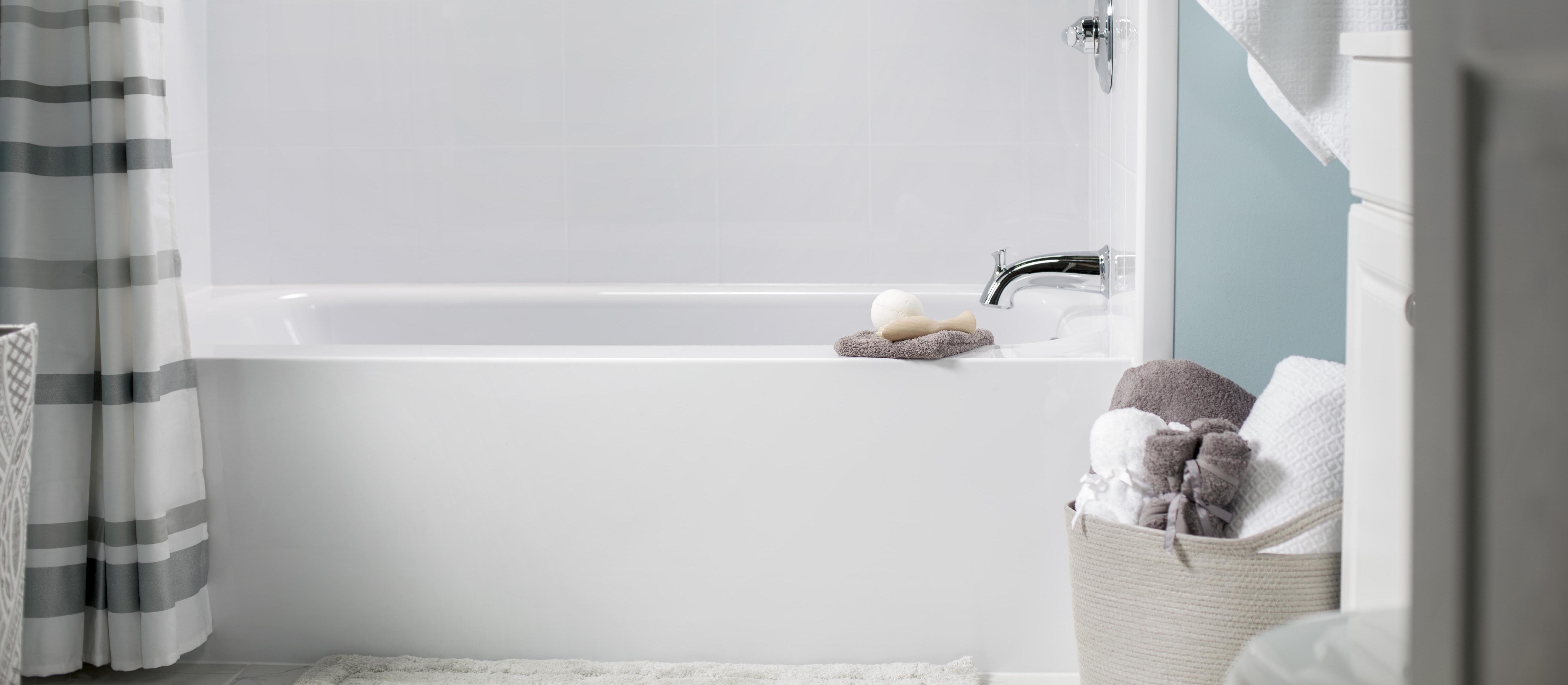 Close-up view of a modern white bathtub with a chrome faucet and built-in corner shelves. The shower curtain features a gray and white striped pattern. A soft towel with soaps rests on the tub’s edge, and a woven basket filled with rolled white and gray towels sits nearby. The space is clean and spa-like with light teal walls and minimalistic decor.