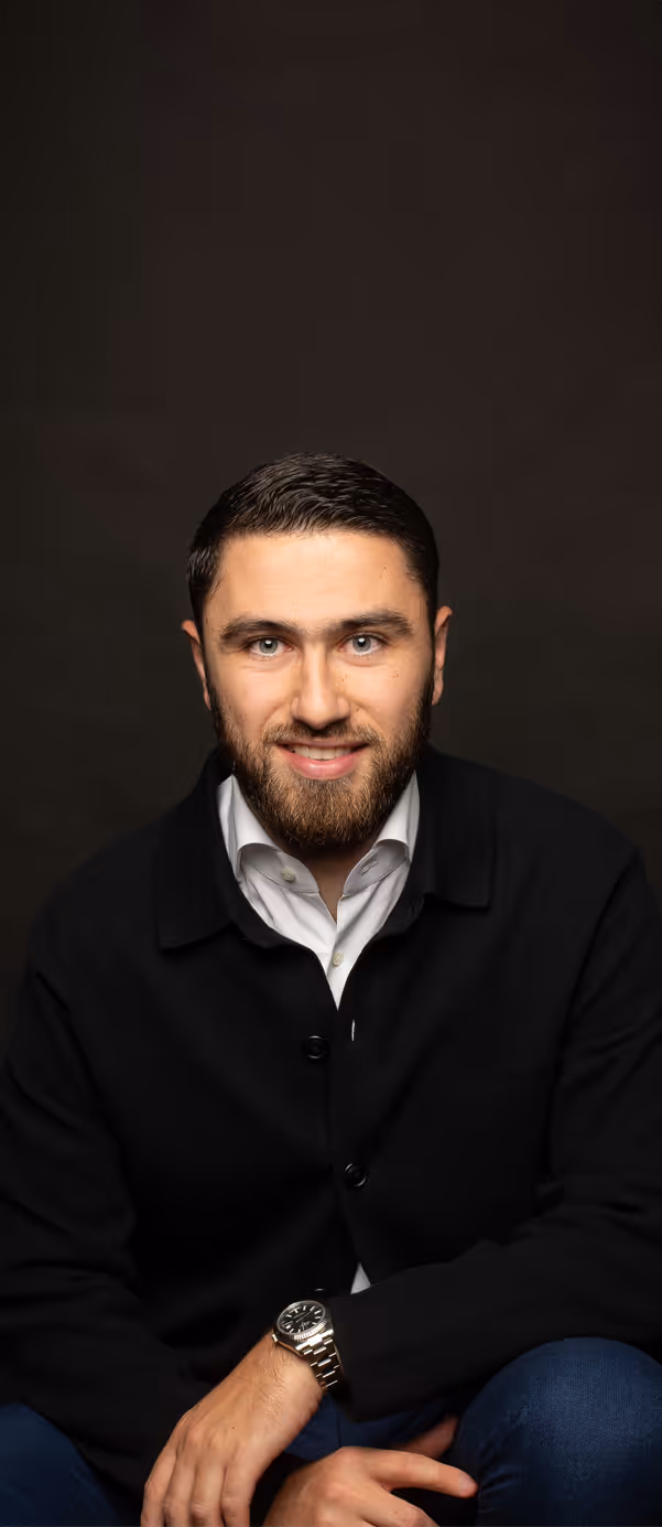 Bearded man wearing a black jacket and white shirt smiling against a dark background.