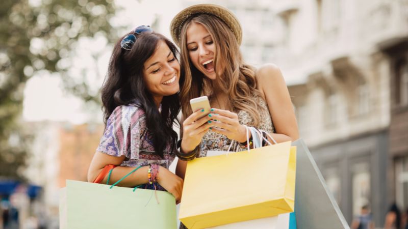 Tow womwn with shopping bags smile at a phone