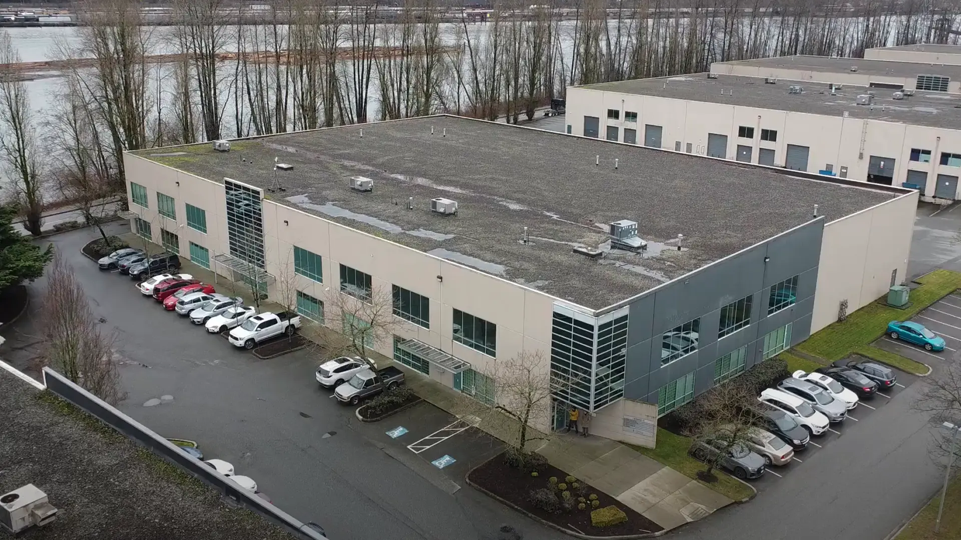 Aerial view of a large commercial building with a flat, gravel-covered roof, surrounded by parked cars and leafless trees near a river.