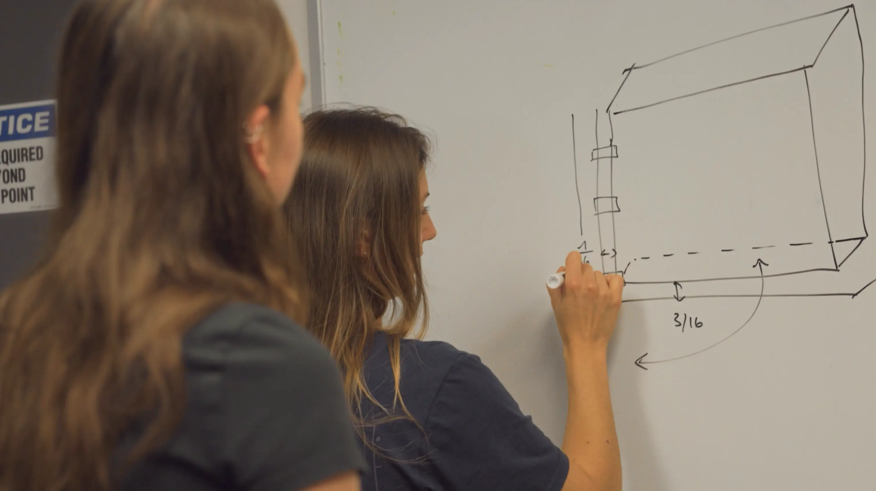 Two women at a whiteboard, one drawing a 3D box diagram with measurements in inches.