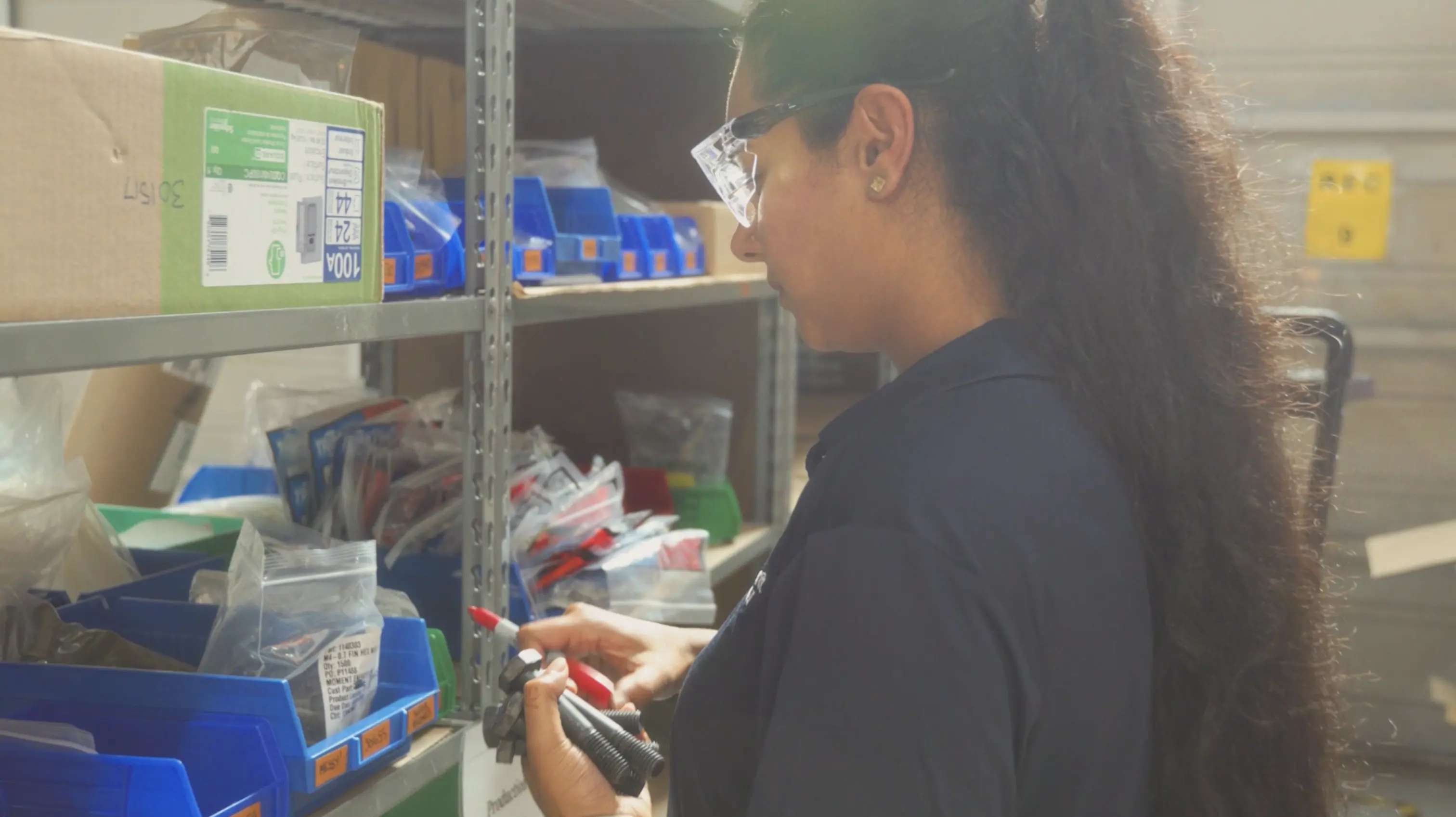 Woman in safety glasses inspecting bolts and screws by metal shelving with blue storage bins in a warehouse.