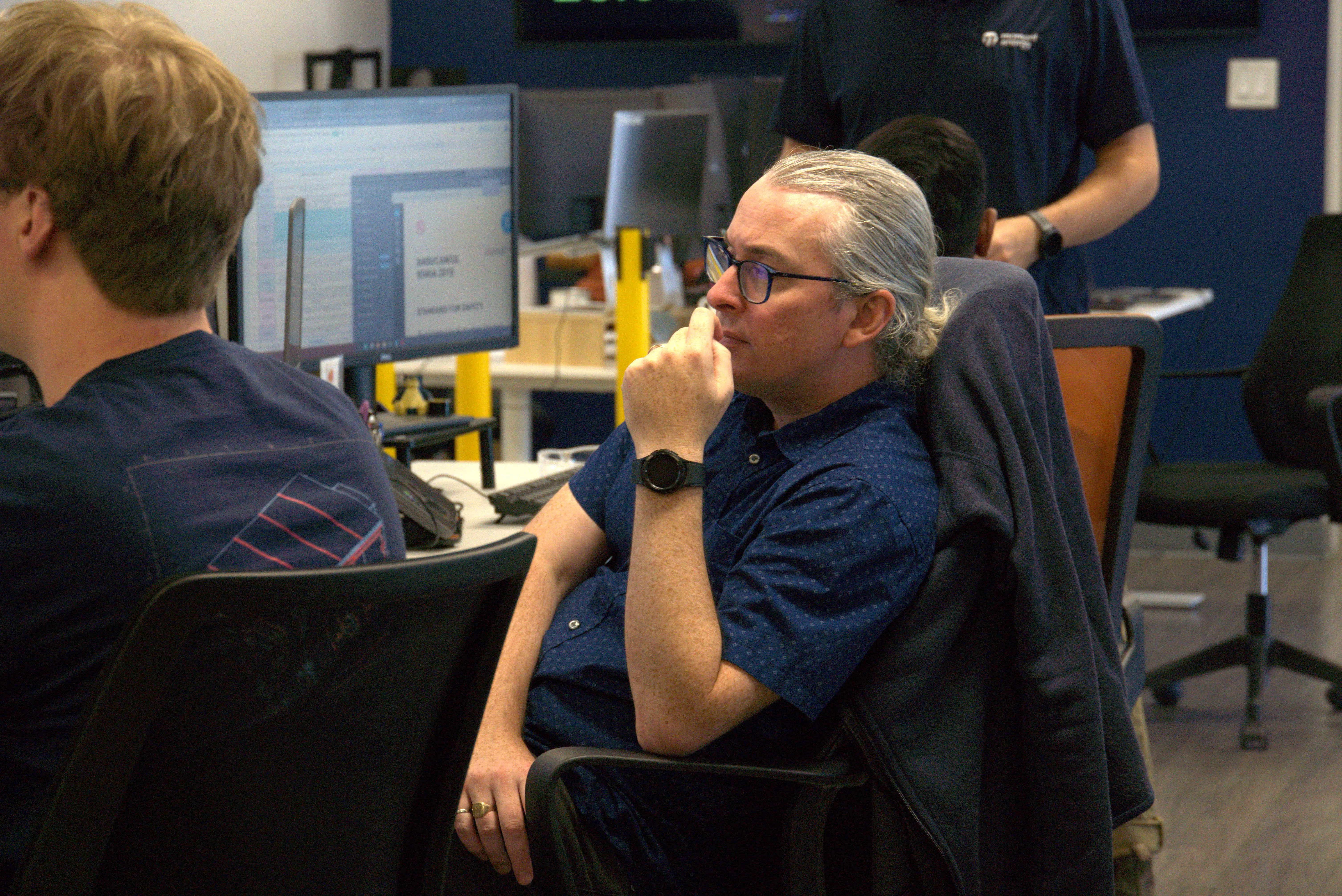 Man with gray hair and glasses thoughtfully sitting in an office chair with a computer screen in the background.