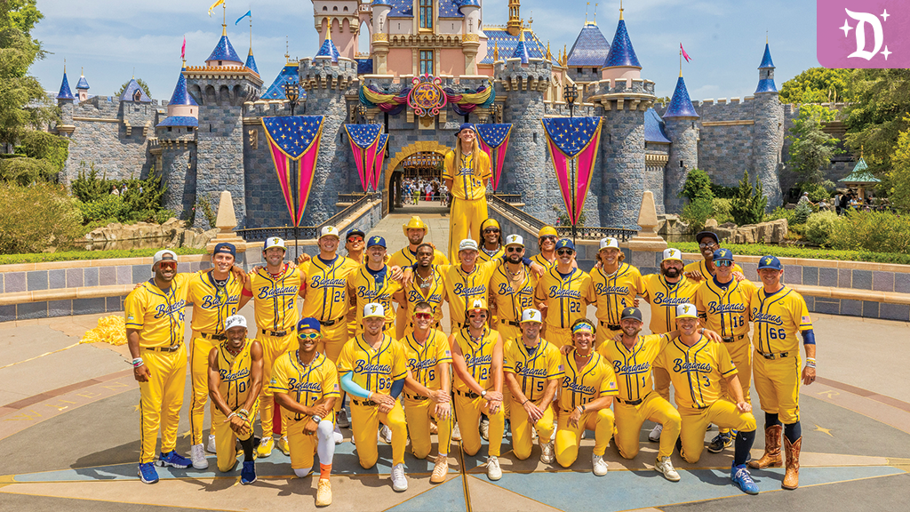 The Savannah Bananas baseball team poses in bright yellow uniforms in front of Sleeping Beauty Castle at Disneyland, with festive red, white, and blue decorations.