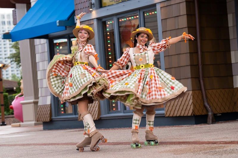 Two women wearing orange and green gingham dresses and straw hats roller-skating and twirling on a pedestrian street in front of a storefront.