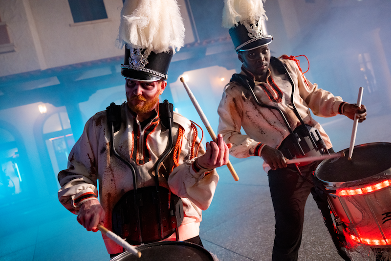 Two marching band drummers wearing tall white feathered hats play red-lit drums on a foggy street under blue lighting