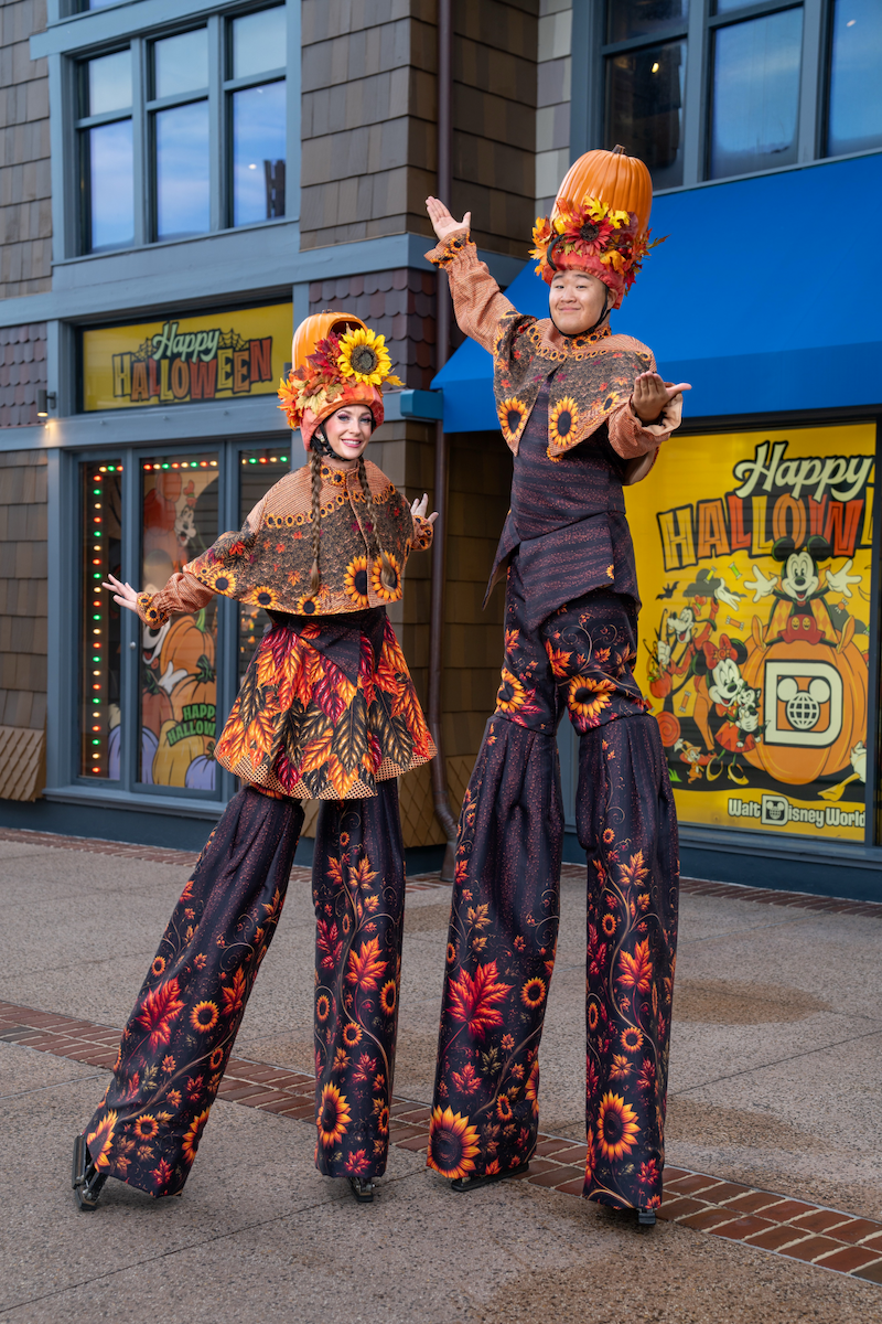 Two stilt walkers in fall costumes with sunflower and leaf prints and pumpkin hats stand before a storefront with Happy Halloween signs.