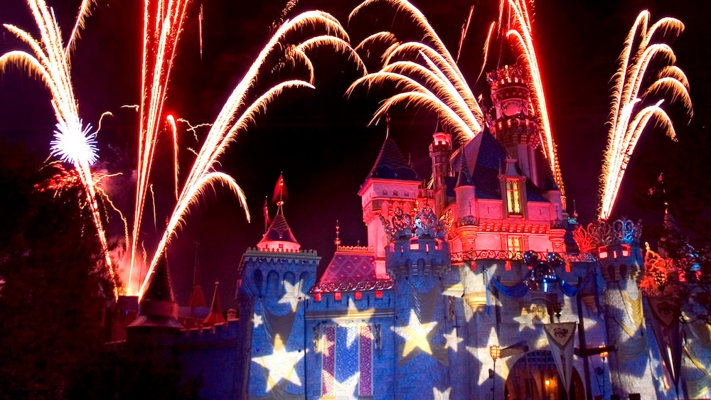 Castle spires and walls lit by colored light and star patterns, with red and white fireworks arcing overhead against a night sky