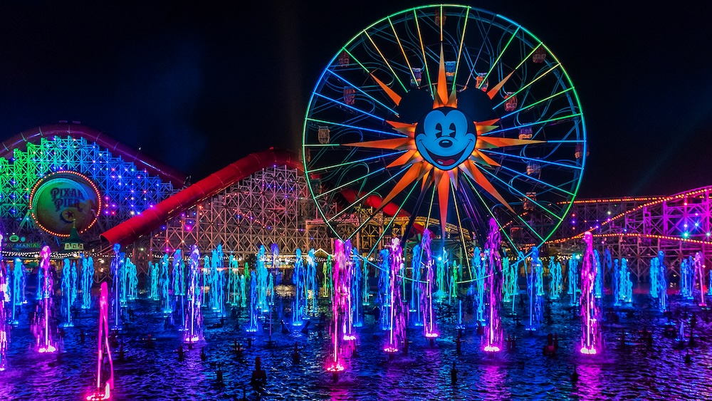 Large illuminated Ferris wheel with Mickey Mouse face behind multicolored dancing water fountains at night in an amusement park