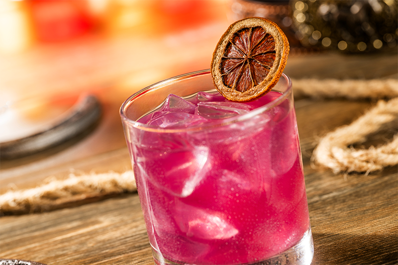 Short clear glass holding pink iced drink, topped with a dried citrus slice on the rim, set on a wooden table.