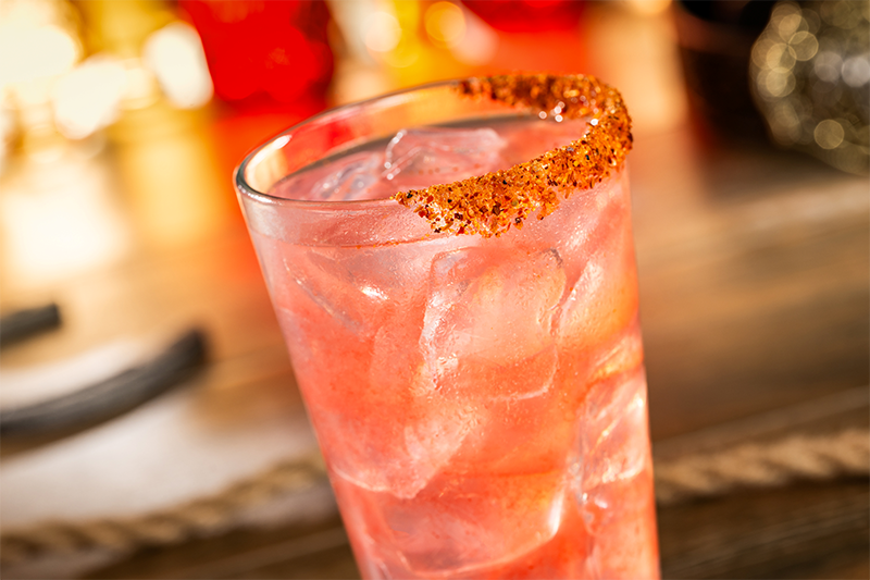 Tall glass filled with a pinkish-red iced drink, rim coated in red chili flakes, blurred bar counter and bottles in background
