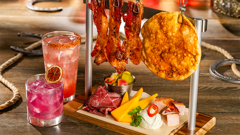 Two pink cocktails with ice and dried citrus garnish beside a rack with hanging bacon and flatbread and a board of sliced roast beef, cheddar and ham