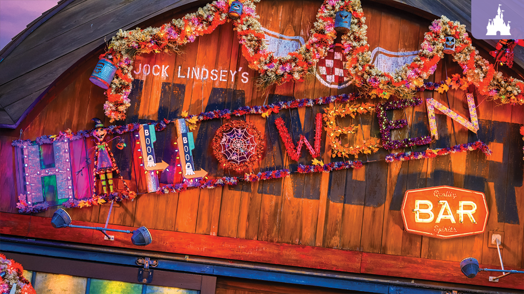 Barn-style wooden bar facade decked in colorful garlands spelling HALLOWEEN above a glowing BAR sign with fall decorations.