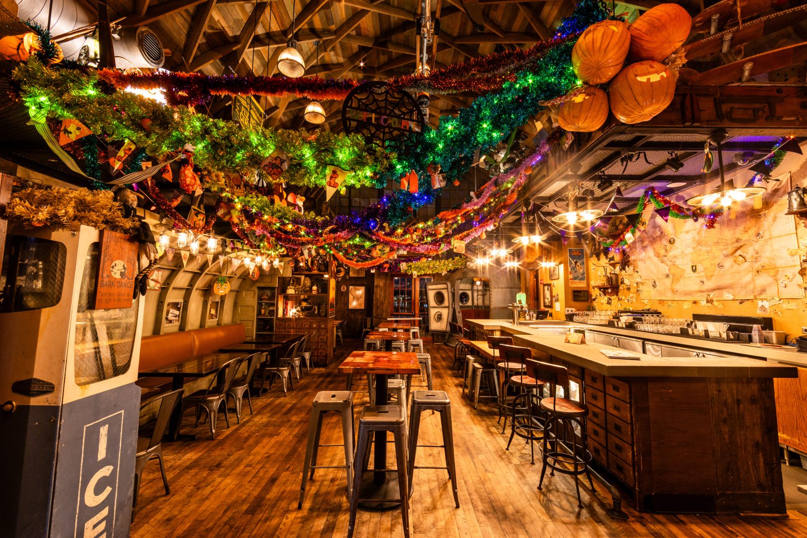 Bar interior decorated with colorful Halloween garlands, carved pumpkins overhead, wooden tables and metal stools lining a wooden floor.