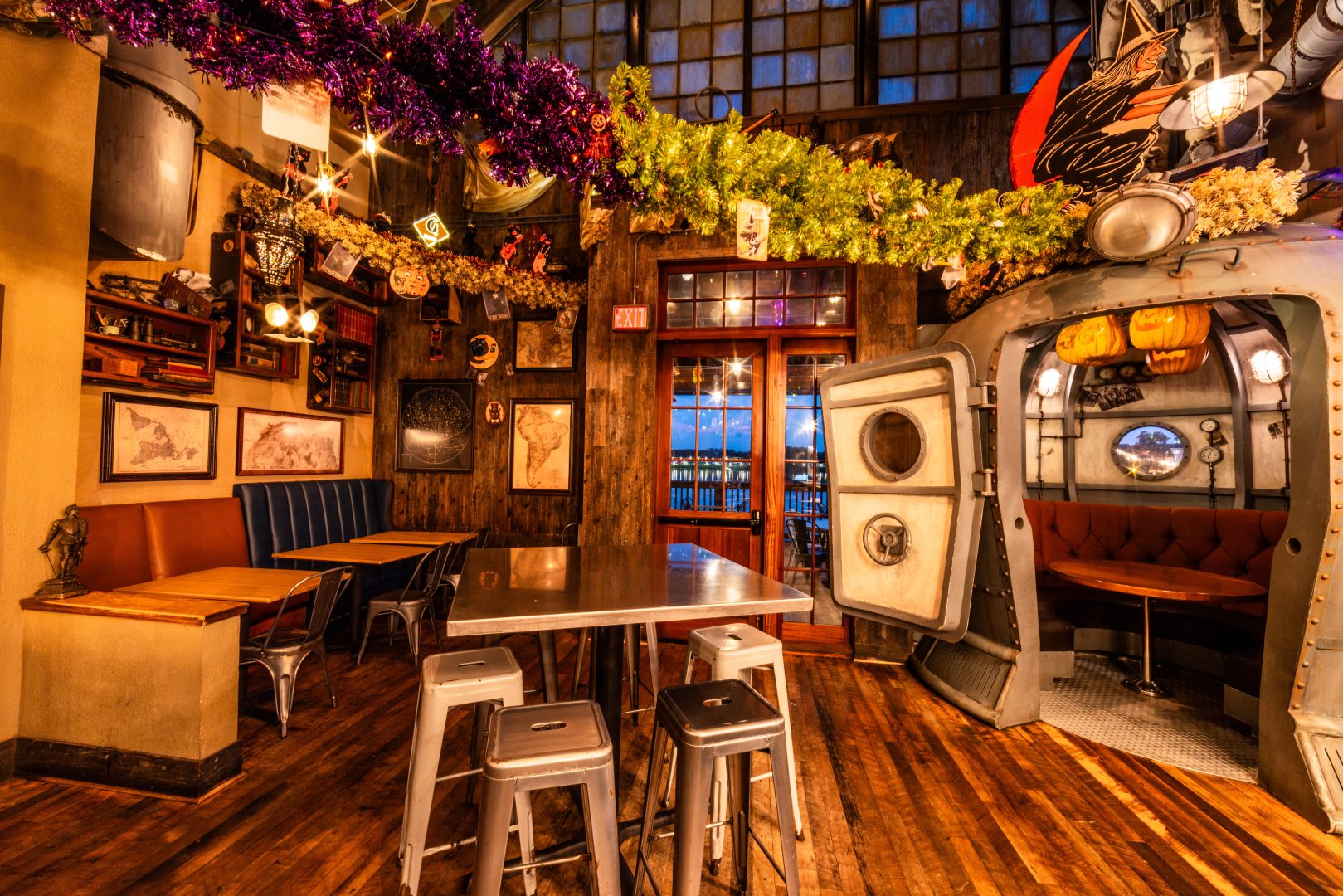 Steampunk-style bar interior with metal tables, stools, a submarine pod booth, wooden floors, maps and Halloween garlands with pumpkin lanterns.