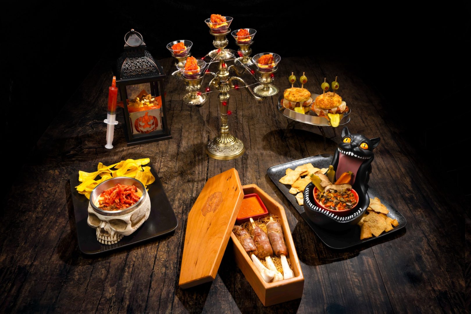 Wooden tabletop with candelabra of dishes, skull bowl with chips, coffin box of skewers with sauce, cat-shaped dip and mini sliders, syringe
