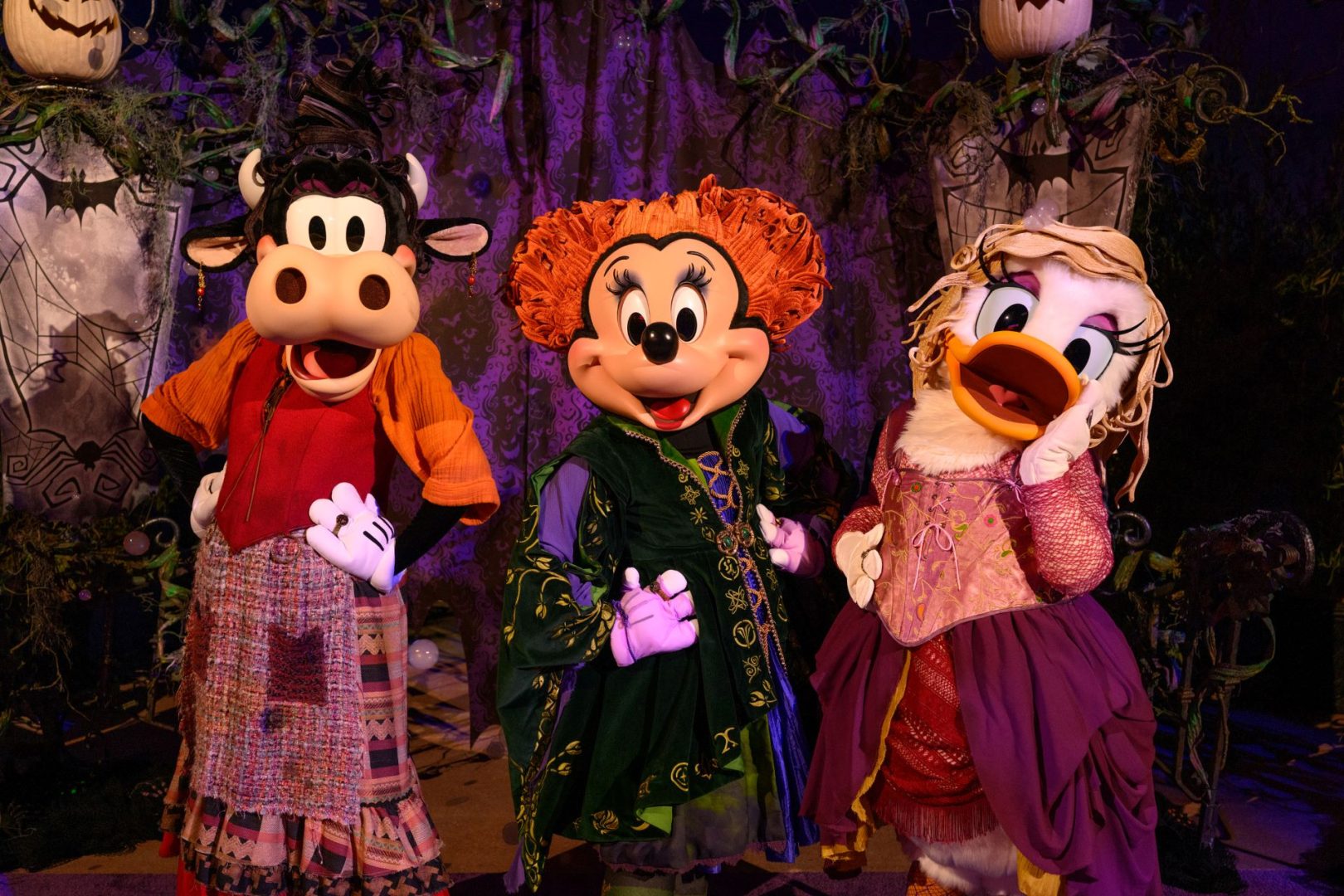 Three costumed Disney characters—a cow, a mouse, and a duck—wearing witch-themed gowns against a purple-spiderweb Halloween backdrop